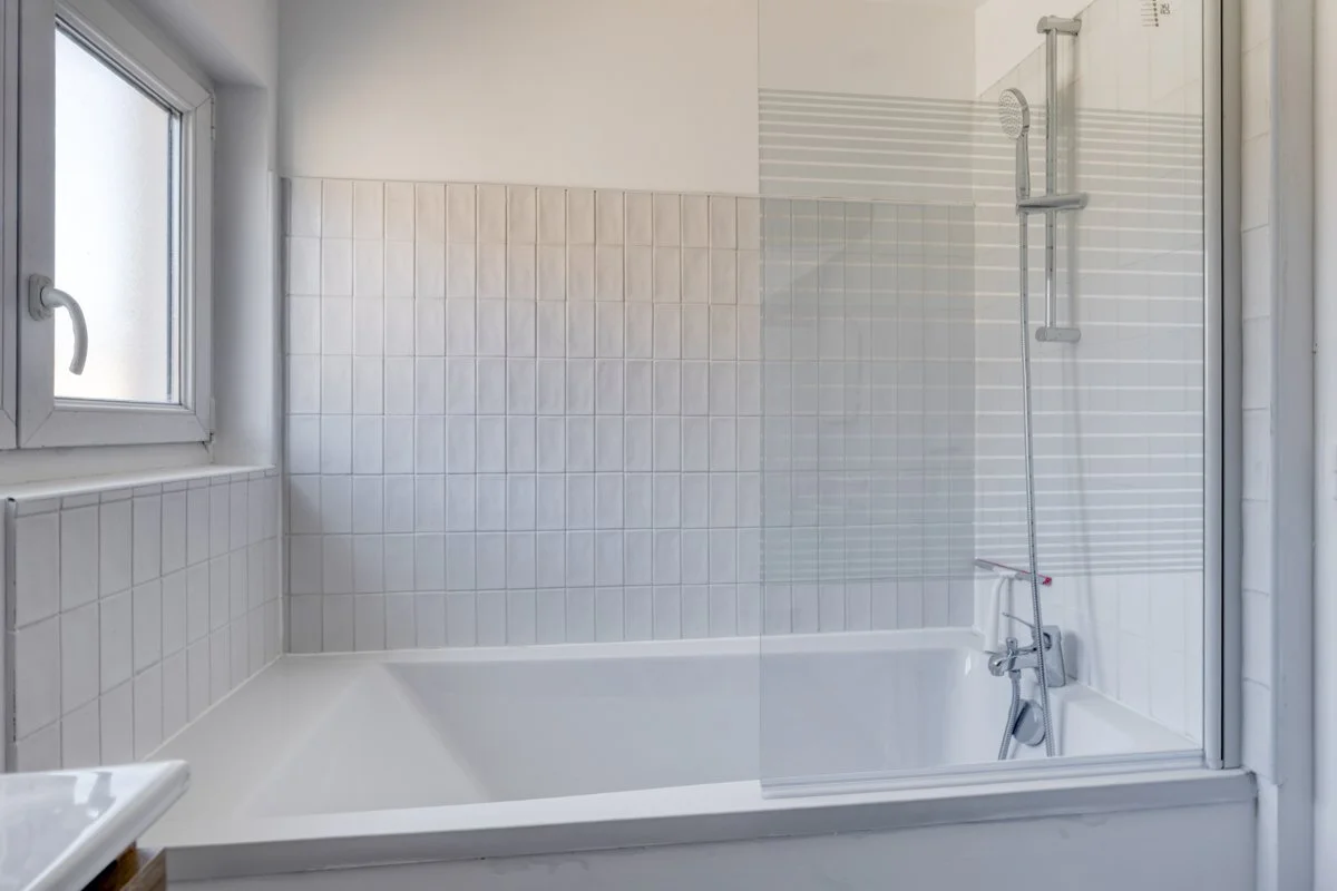 Salle de bain avec baignoire, mur en carreaux blancs, fenêtre avec poignée, cabine de douche avec paroi en verre et poignée, pomme de douche.