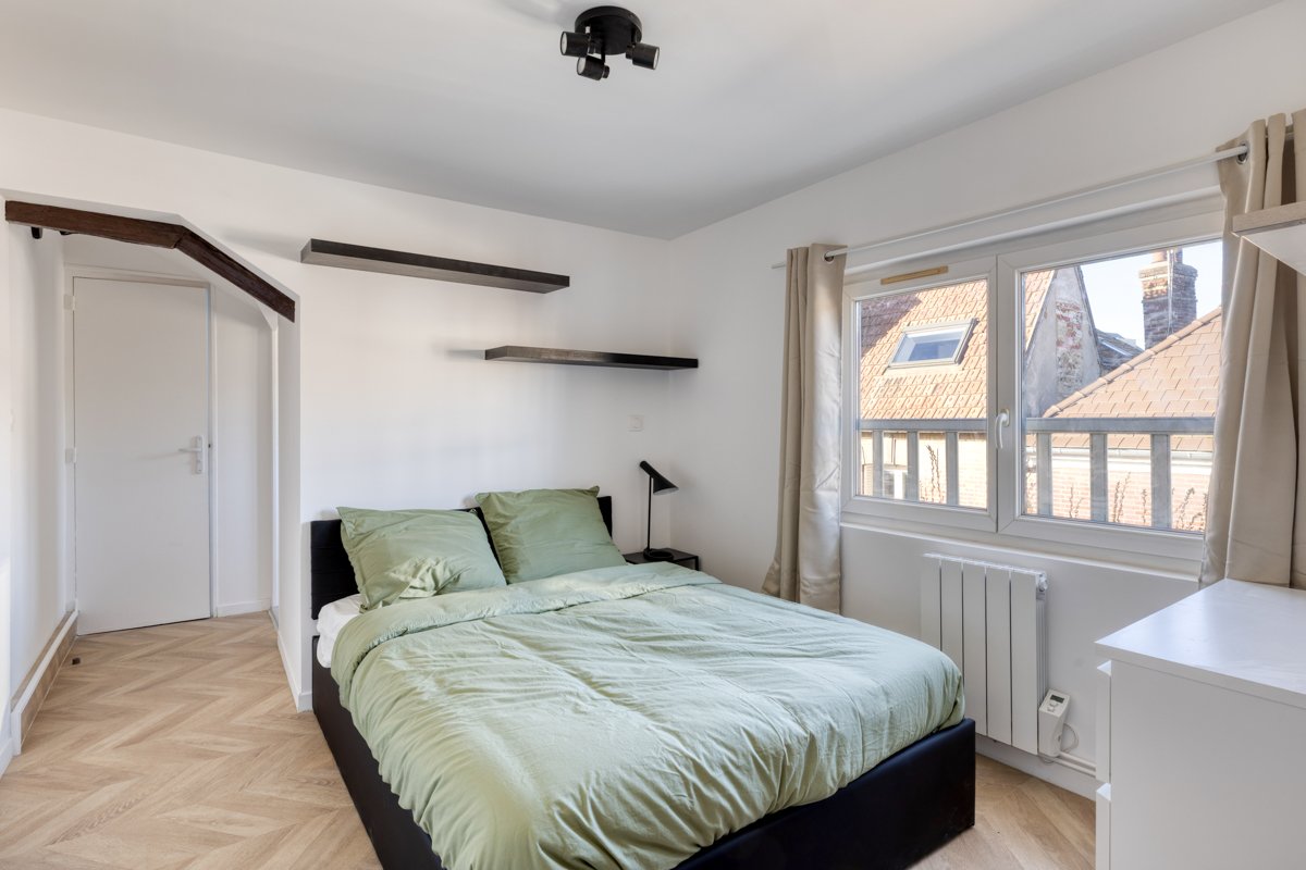 Chambre avec lit en vert, deux étagères noires, fenêtre laissant entrer lumière, rideaux beiges, plafond avec éclairage, plancher en bois.