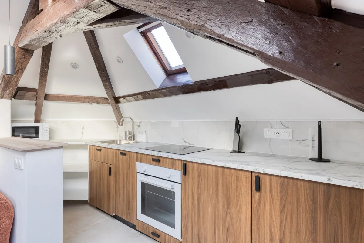 Cuisine avec poutres en bois apparentes, fenêtre de toit, plan de travail en marbre, four encastré, micro-ondes, et prises électriques.