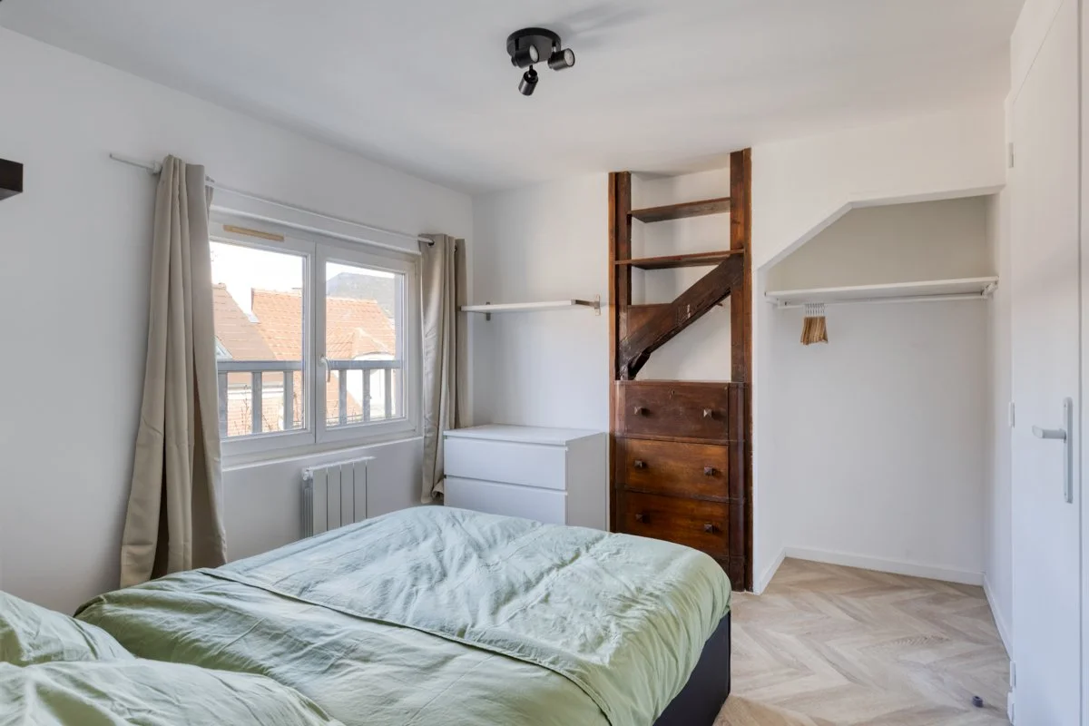 Chambre lumineuse avec deux lits, fenêtre avec rideaux, et une cloison en bois avec des étagères, un tiroir en bois foncé, un commode blanche, et un rayonnage blanc derrière la porte.