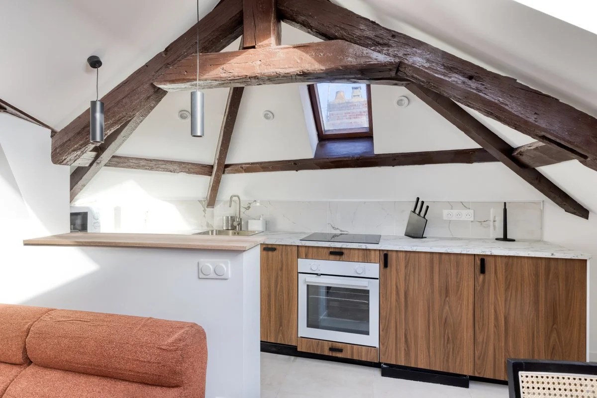 Cuisine dans un loft sous un plafond en pente avec poutres en bois apparentes, petite fenêtre de toit, et un comptoir en marbre avec des appareils électroménagers en bois