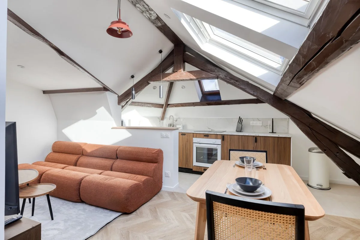 Salon et cuisine dans un appartement avec poutres en bois apparentes et lucarnes au plafond, meubles modernes, sofa orange, table à manger en bois, vaisselle