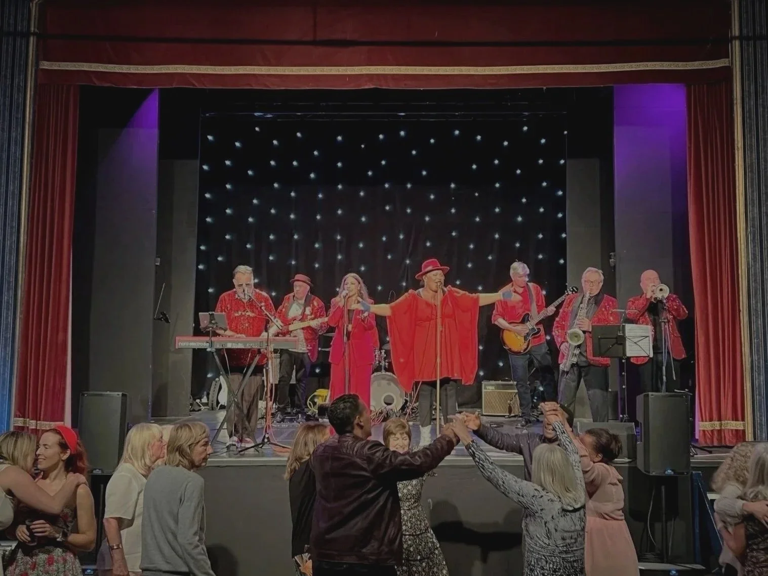 Band performing on stage with audience dancing in front, all dressed in red, in an theater with red curtains and starry backdrop.