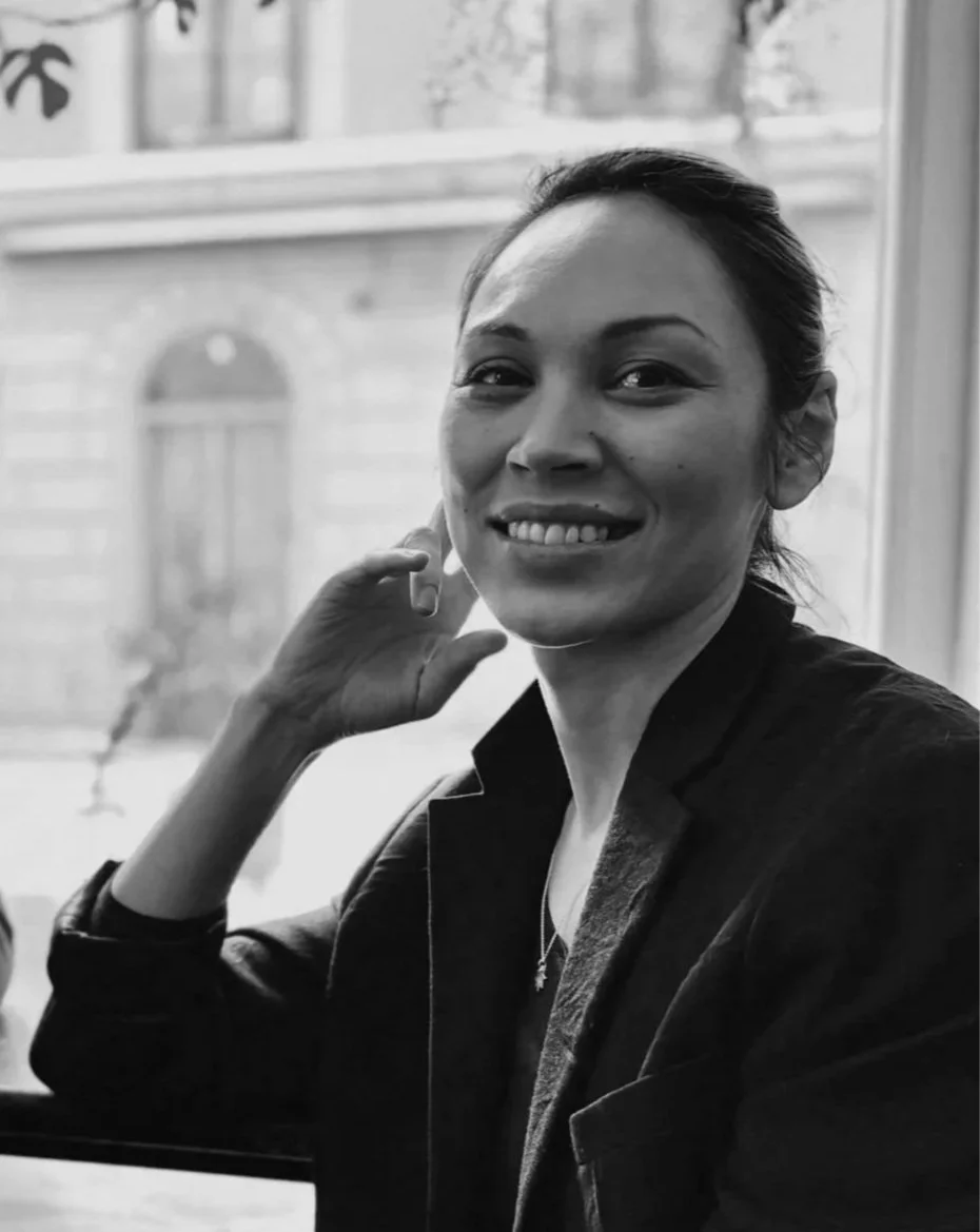 Black and white photo of a smiling woman sitting by a window, with her hand resting near her face, indoors, in natural light.