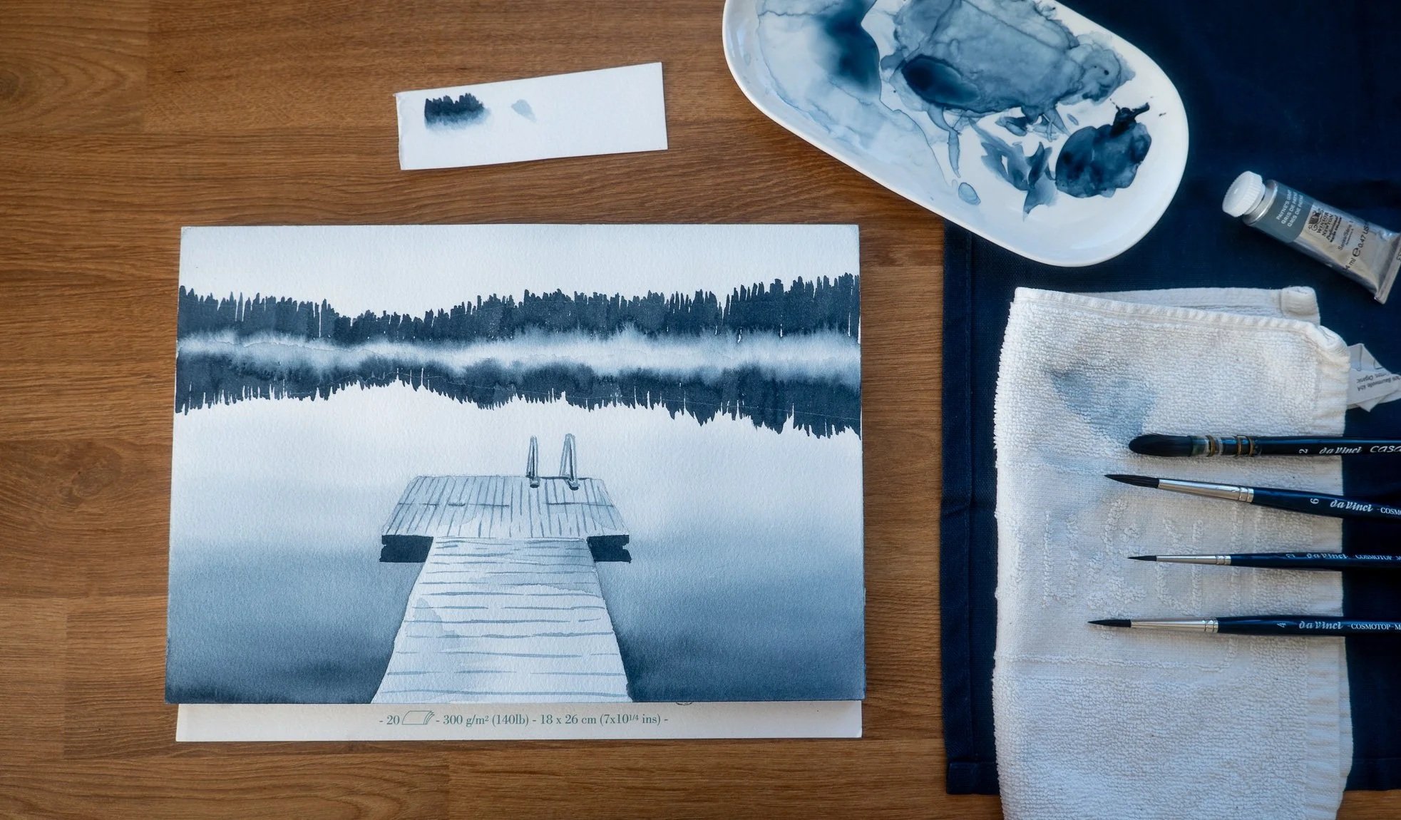 A watercolor painting of a lake with a wooden dock leading into the water, with a forested shoreline and mountains in the background. Art supplies including brushes, a palette, and a cloth are arranged around the painting.