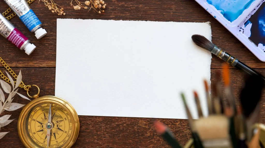 Empty white sheet of paper surrounded by watercolor paints, paintbrush, tubes of paint, a gold compass, and art supplies on a wooden surface.