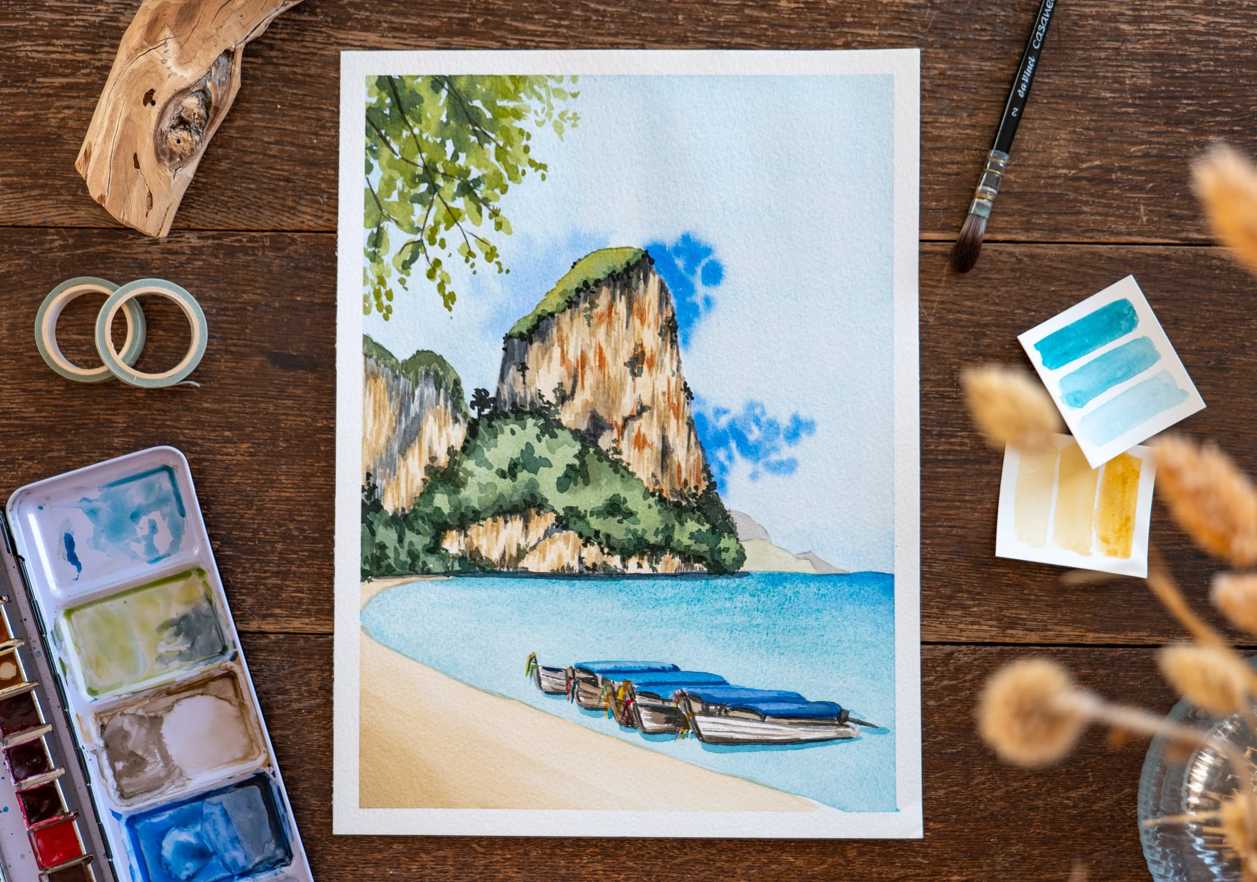 A watercolor painting of a coastal landscape with cliffs, a beach, boats, and a partly cloudy sky, surrounded by painting supplies on a wooden table.