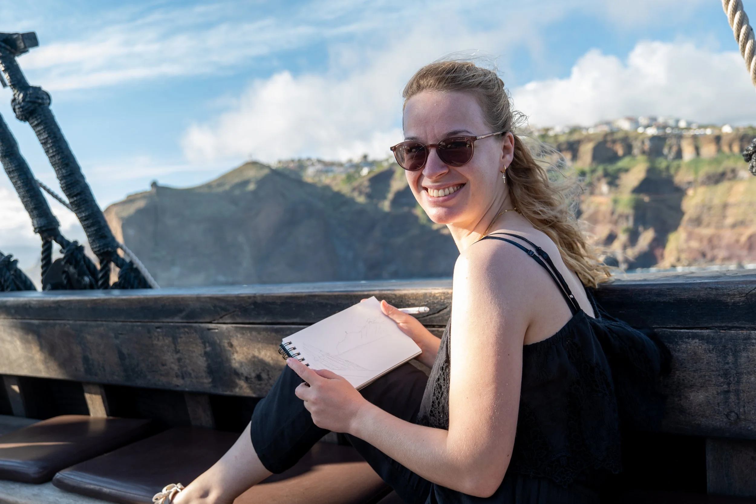 A woman wearing sunglasses sitting on a boat with a sketchbook in her lap, smiling at the camera, with cliffs and a partly cloudy sky in the background.