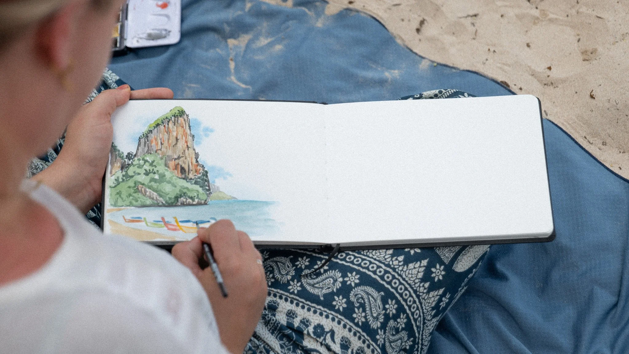 A person sitting on a beach, sketching a landscape scene of a large rocky hill with green foliage, a sandy beach, and colorful kayaks on the water.