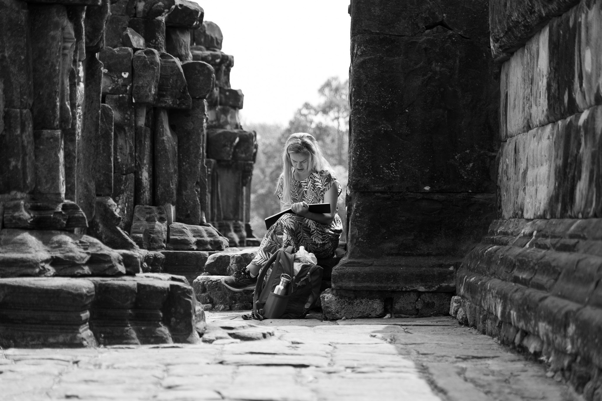 A woman sitting on the ground between ancient stone ruins, reading a book or map, with a backpack beside her.