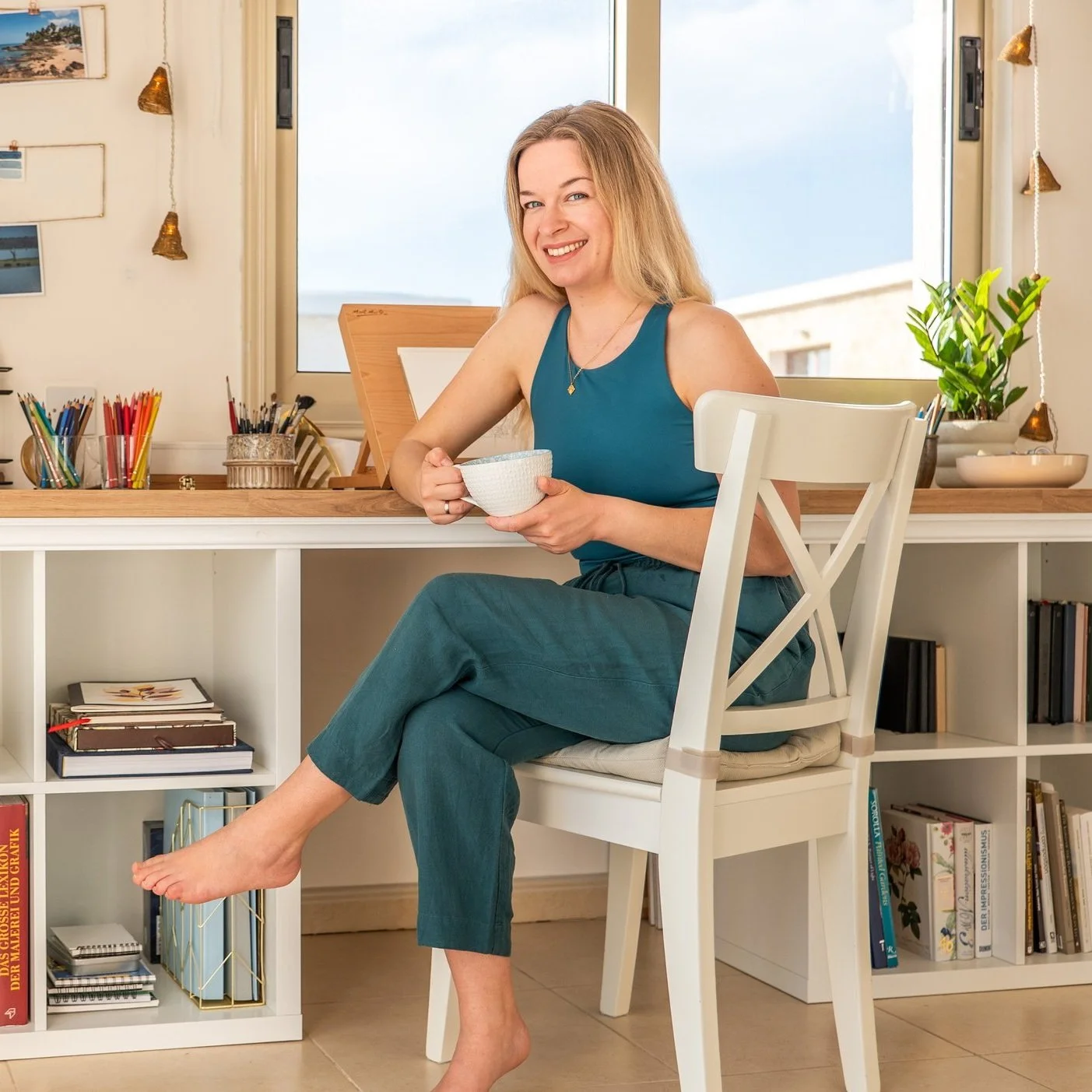 A smiling woman with long, blonde hair sitting at a white desk, holding a white bowl, in a room with a window showing a bright sky outside. The desk has books, colorful pens, and plants on it.
