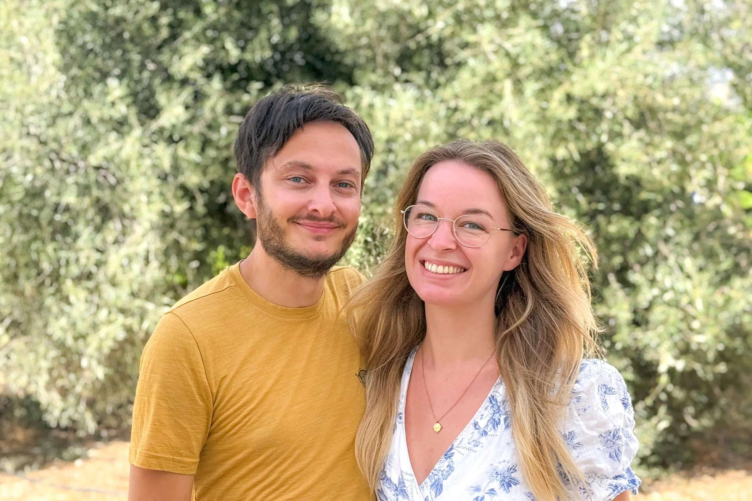 A smiling couple standing outdoors in front of trees, with the man wearing a yellow T-shirt and the woman wearing glasses and a white blouse with blue floral patterns.