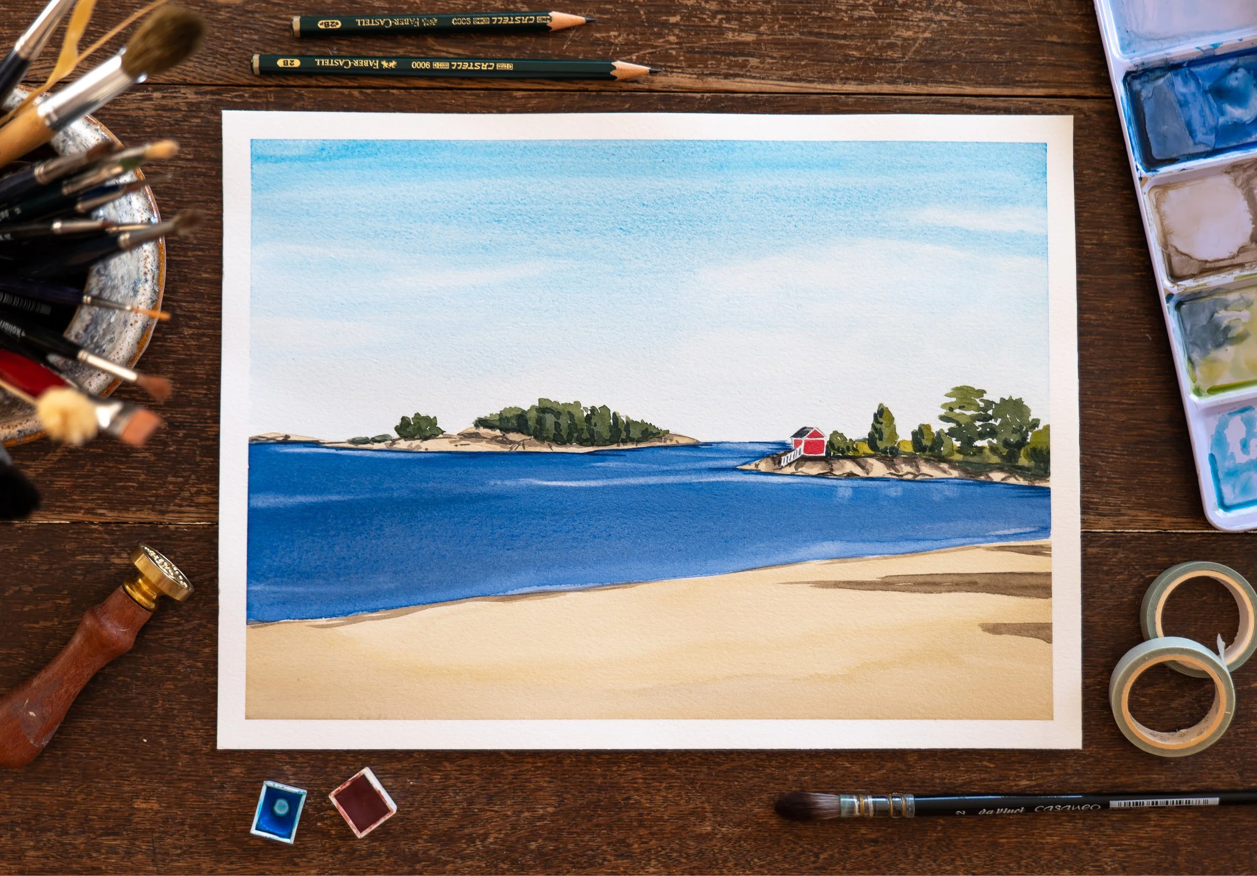 A watercolor painting of a beach scene with blue water, sandy shore, and green trees, with a red building in the distance, surrounded by art supplies on a wooden surface.
