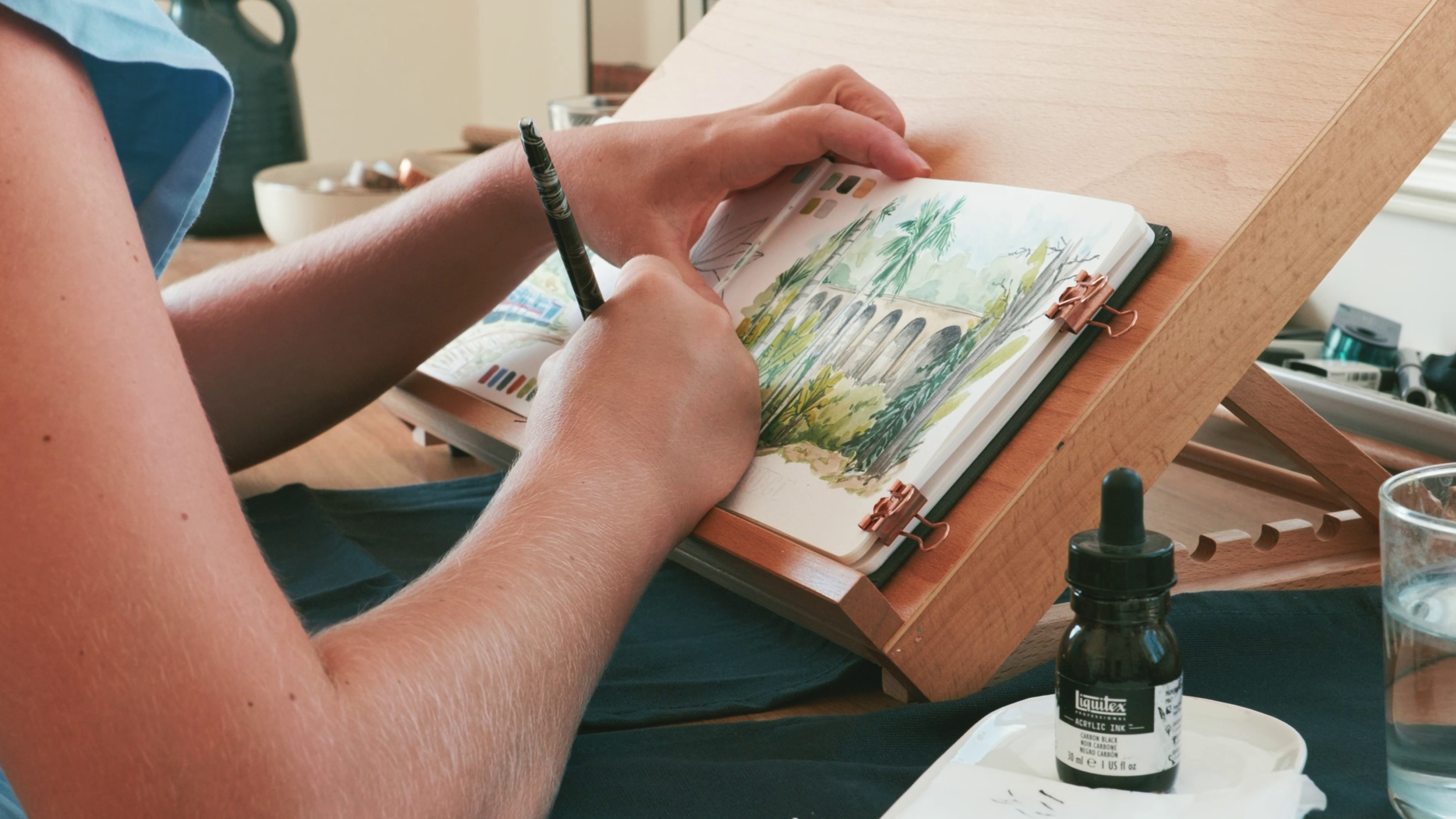 Person painting a watercolor landscape of a bridge, arches, and trees on a sketchbook placed on a wooden artist's easel.