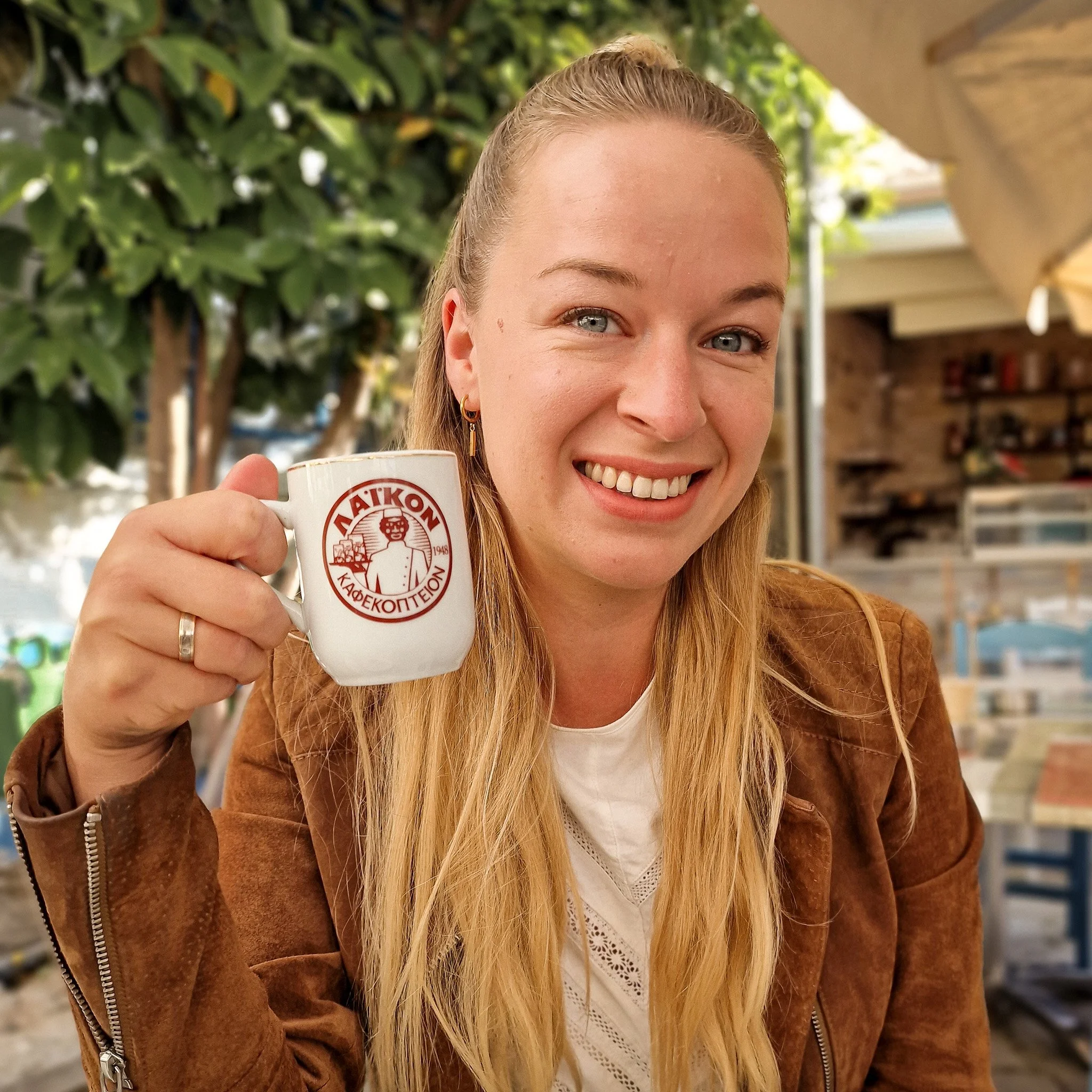 A woman with long blonde hair smiling and holding a coffee mug with Greek writing, sitting outdoors at a café with green foliage and a brick wall in the background.