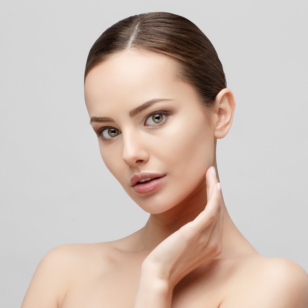 Close-up of a young woman with fair skin and brown hair, gently touching her face with her hand, against a plain light gray background.