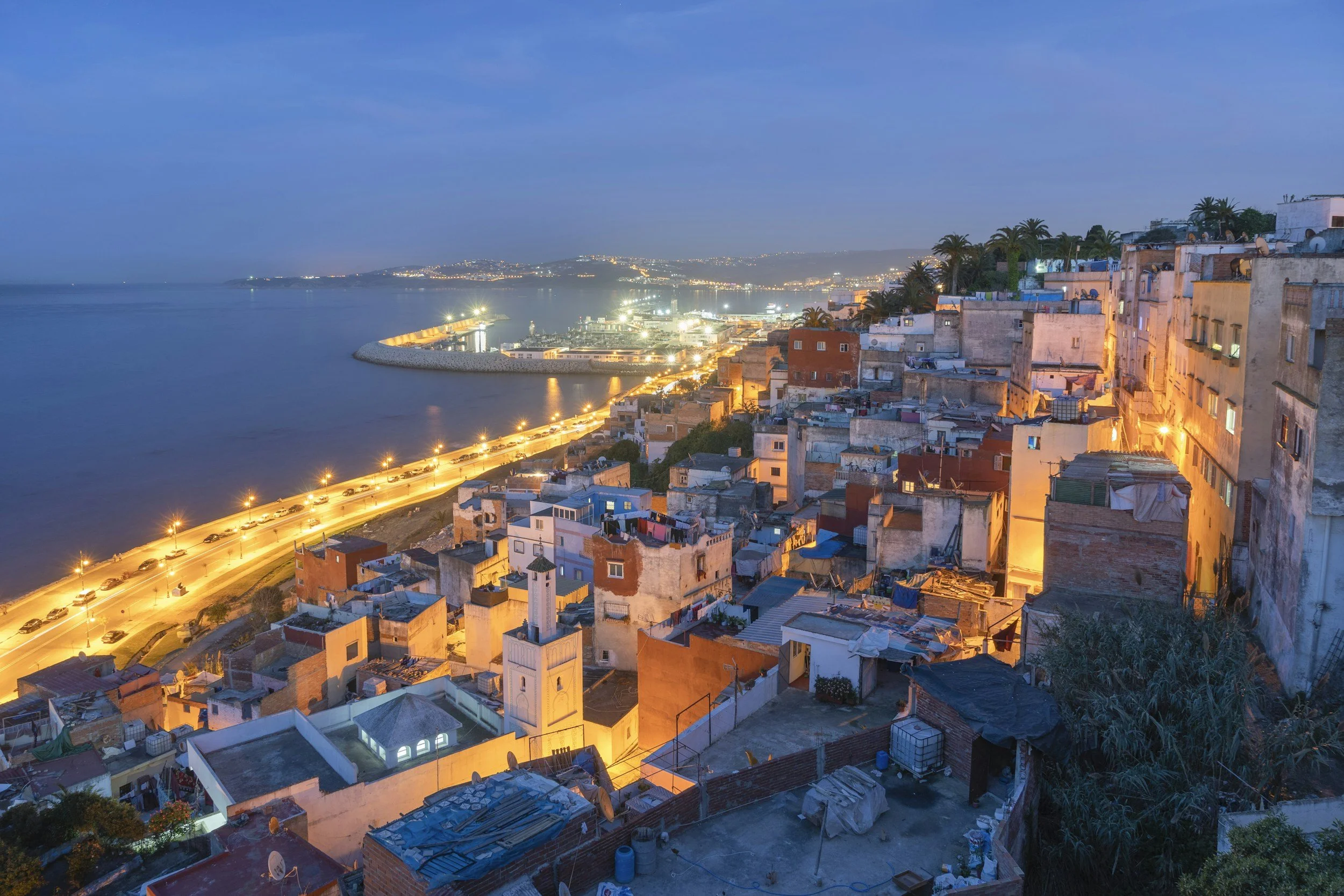 Cityscape of houses along a coast at dusk, with a lit street and water in the background.