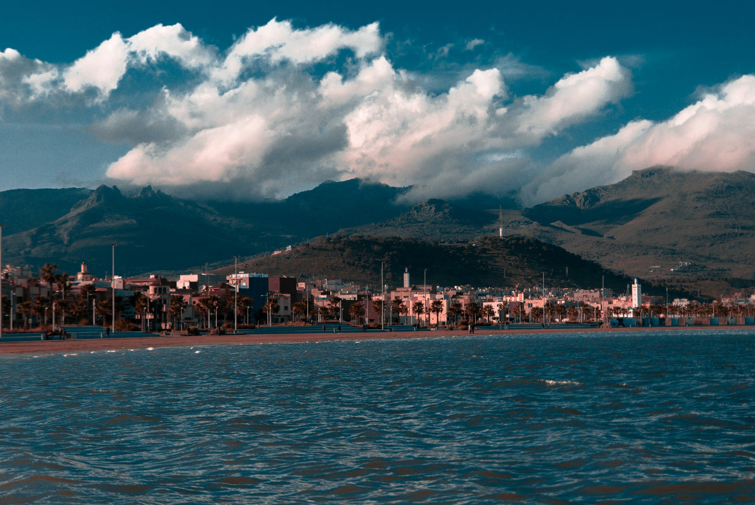 View of a cityscape near water with mountains in the background and partly cloudy sky.