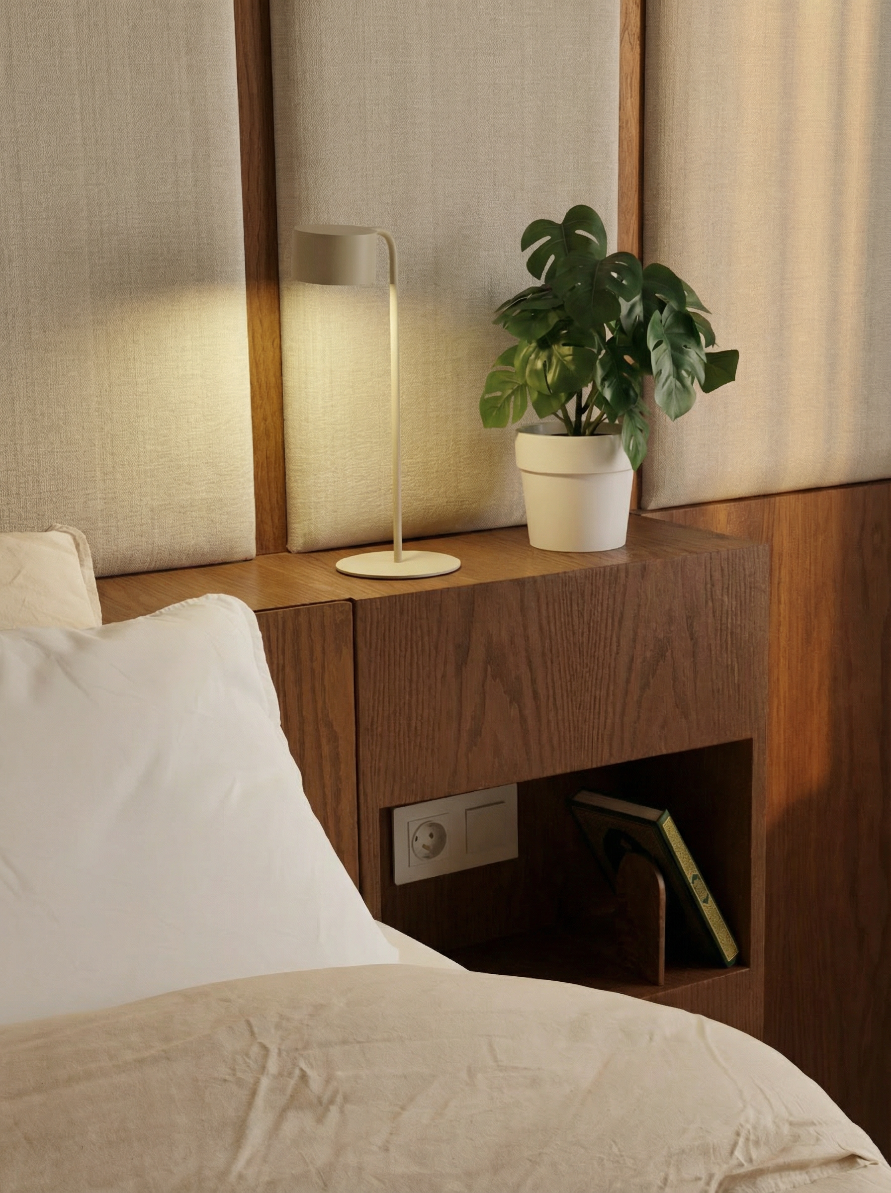 A bedroom interior with a bed, white pillow, beige bedsheet, wooden headboard, bedside table with a small white lamp, potted plant, wall socket, and a book on a stand.