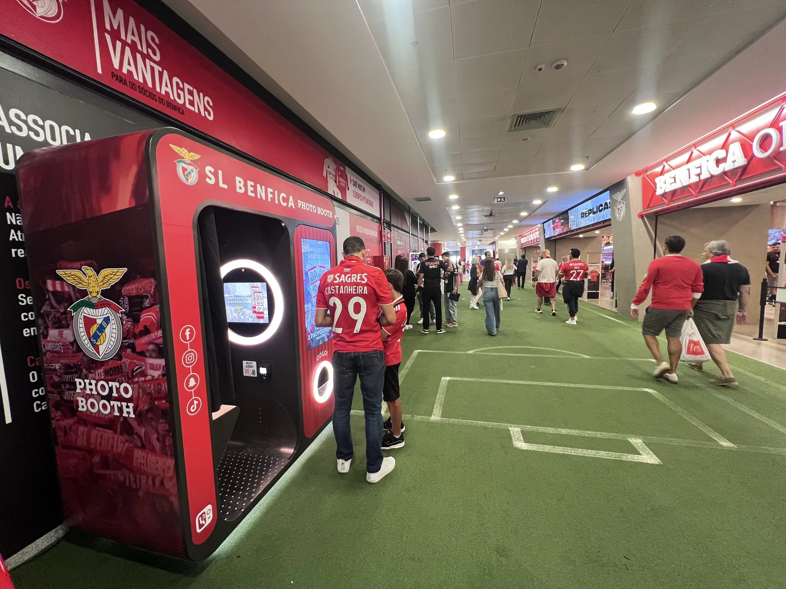 People walking through a soccer fan shop area with Benfica branding, including a photo booth, souvenir displays, and fans in Benfica jerseys.
