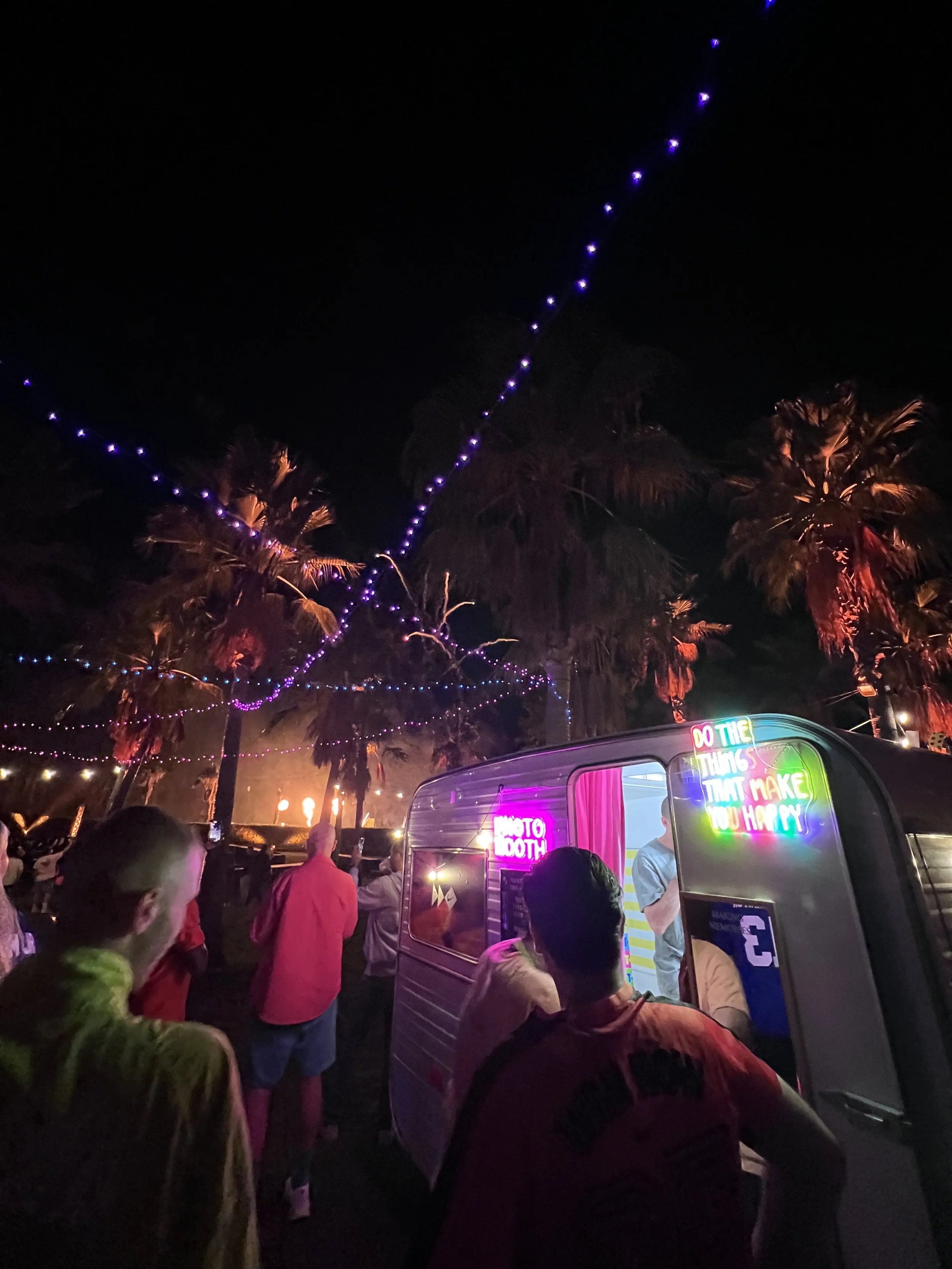 Night scene at a carnival or fair with string lights and palm trees, a neon sign on a food truck or photo booth, and people waiting in line.