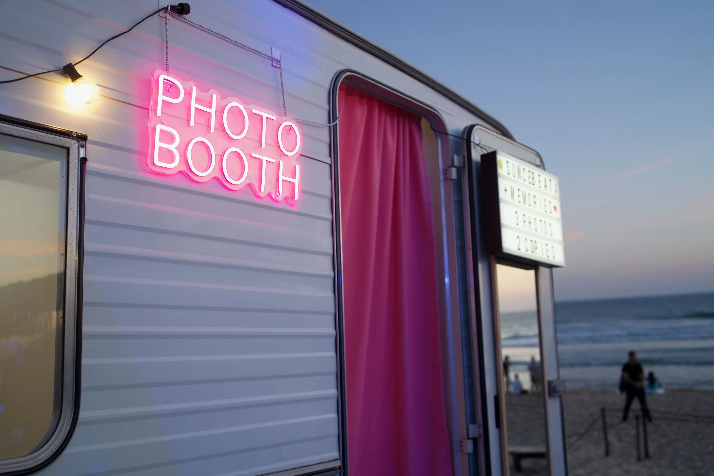 A mobile photo booth trailer on the beach at sunset, with a pink neon sign that reads 'PHOTO BOOTH', pink curtains inside the entrance, and a signboard listing services in the background, with people on the beach and the ocean in the distance.