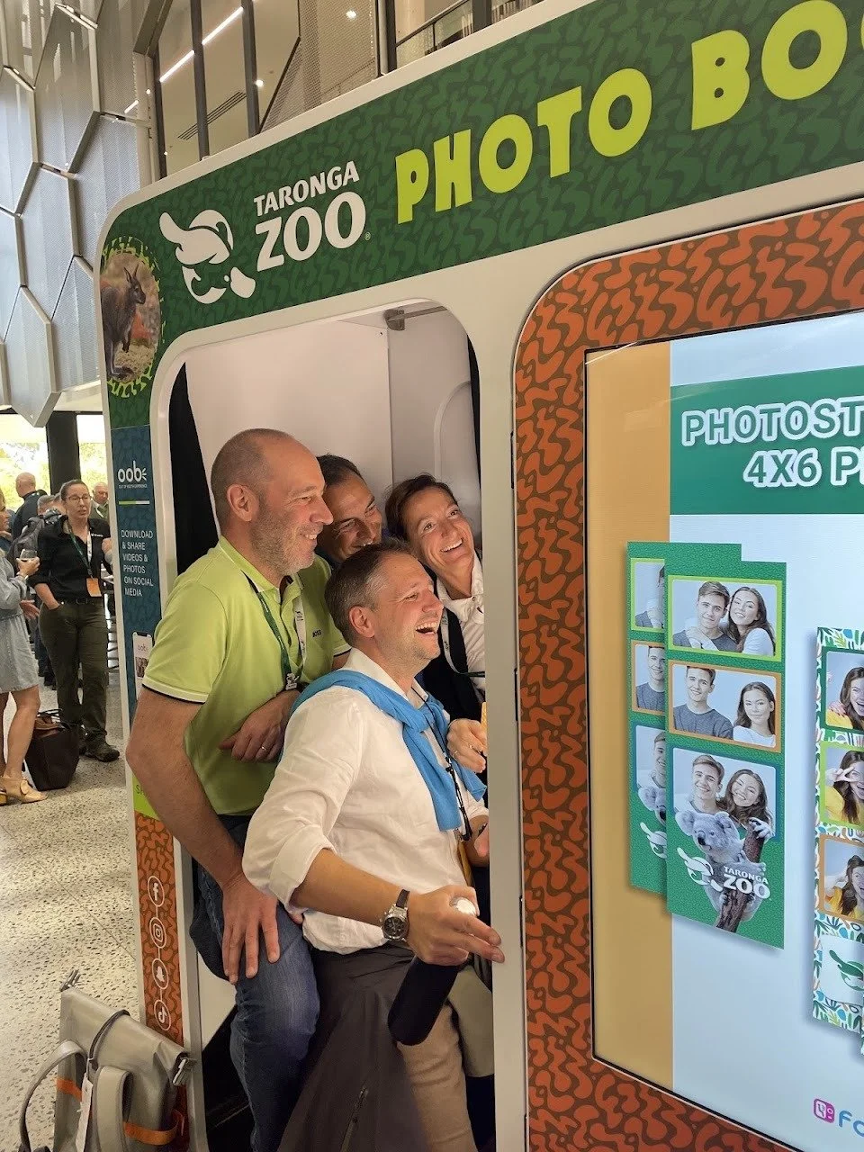 Group of four people taking a fun photo in a photo booth at Taronga Zoo, with the booth's colorful signage visible.
