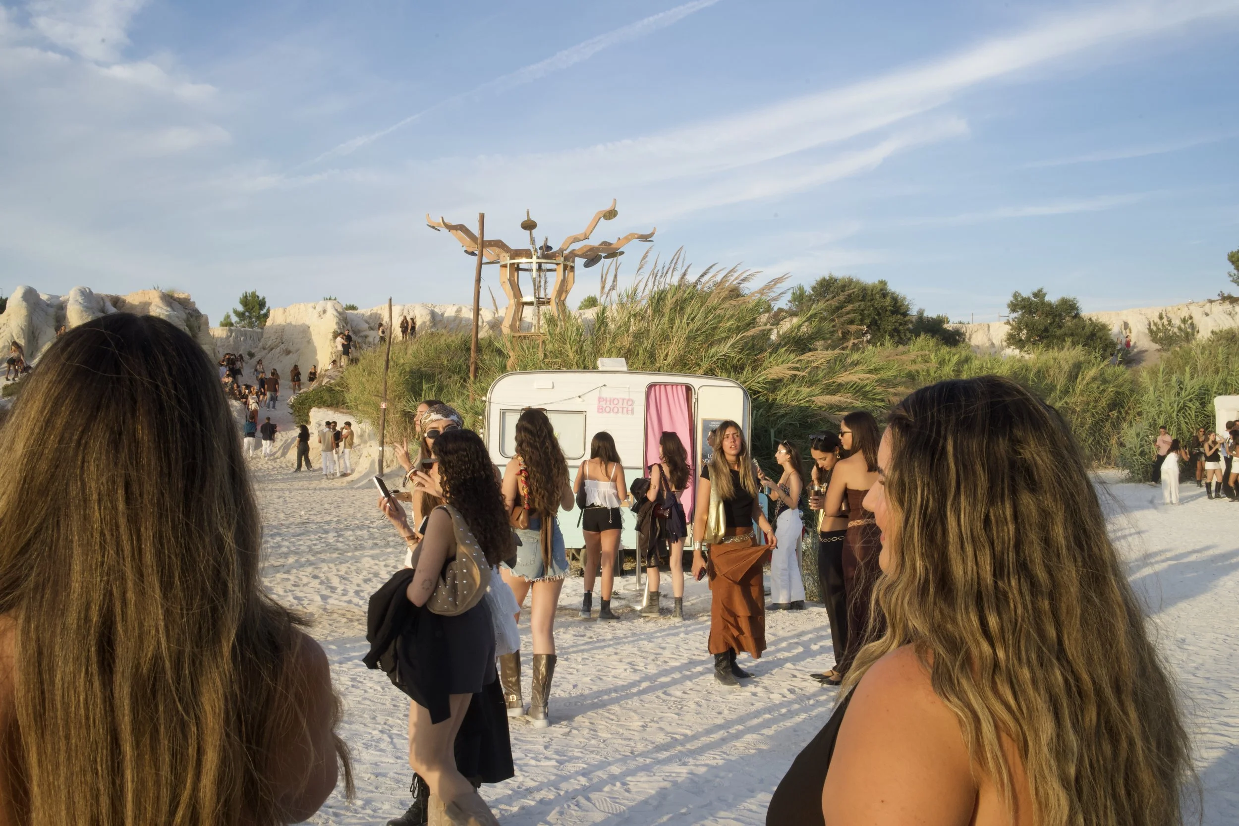 Group of people at an outdoor event near a photo booth, with a desert landscape and a sculpture resembling a tree or branch structure in the background.