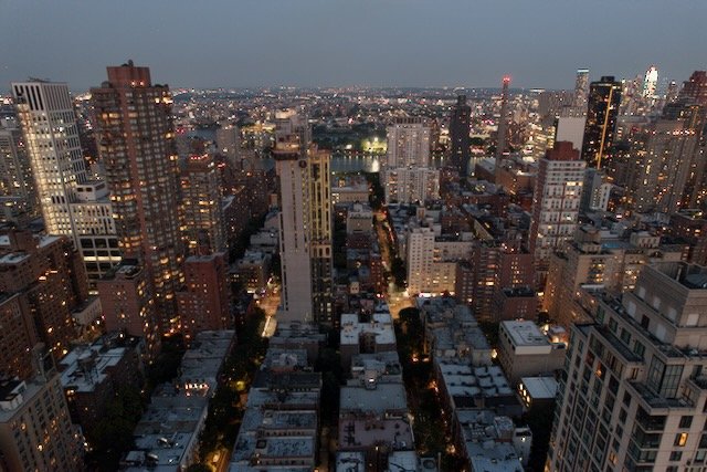 Aerial view of a cityscape during early evening with illuminated skyscrapers and residential buildings, streets visible below.