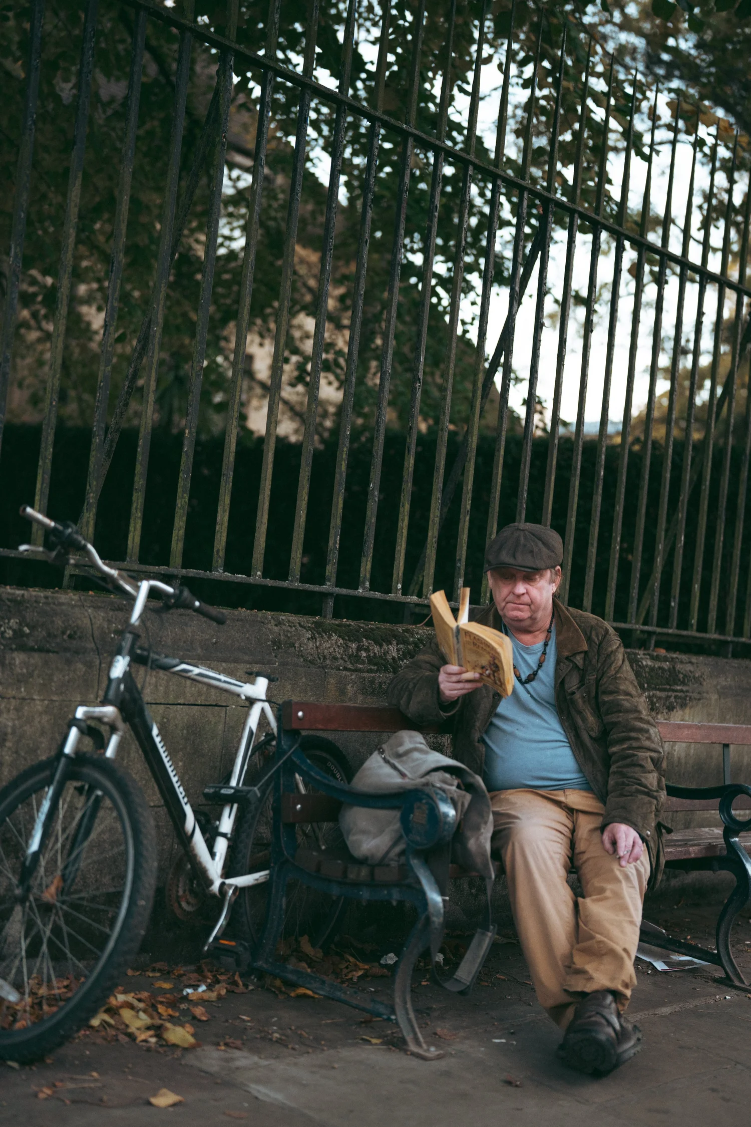 A man with a bicycle leaning against railings, holding up a book and looking directly at the camera.