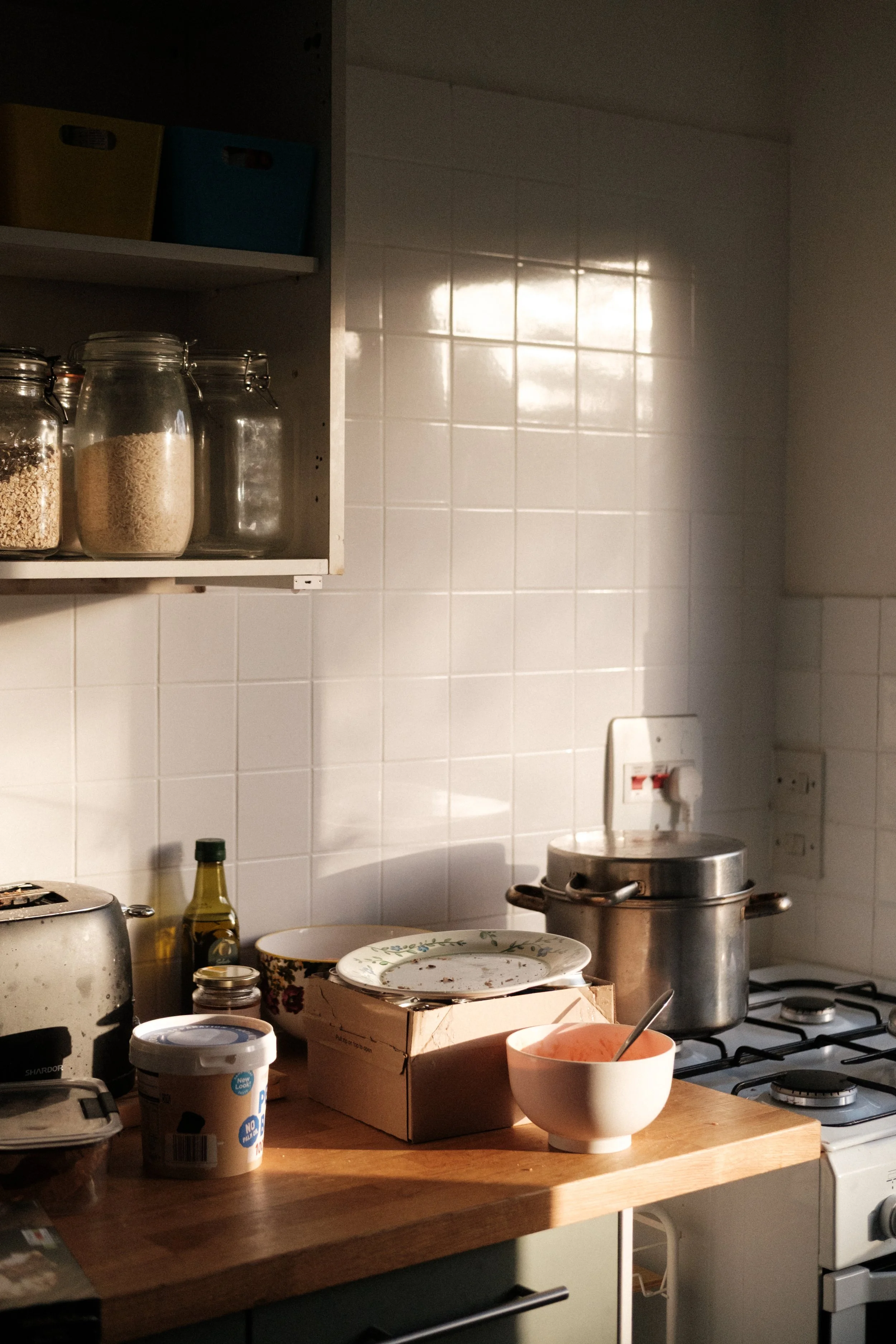 A cluttered kitchen countertop with various kitchen items including a bowl, a pizza box, jars, a toaster, a bottle, and a pot on the stove. A tiled wall is in the background with a light reflection.