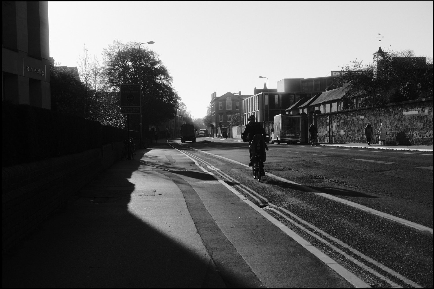 Black and white photo of a cyclist silhouetted against morning light on a wide street beside a stone wall, with a clock tower in the distance.