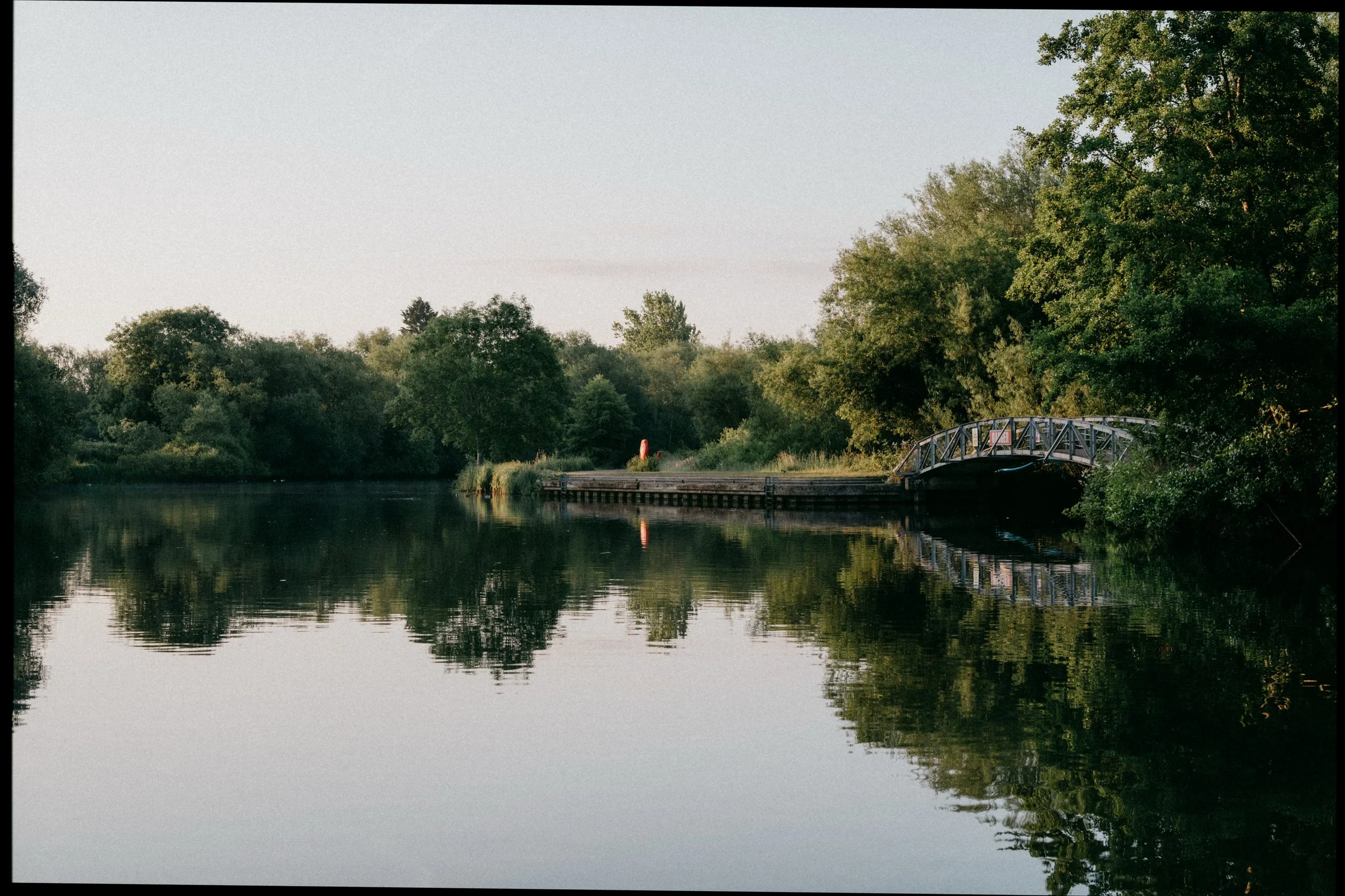 A calm river reflecting green trees on both sides, with a small metal bridge on the right side and a wooden dock extending into the water.