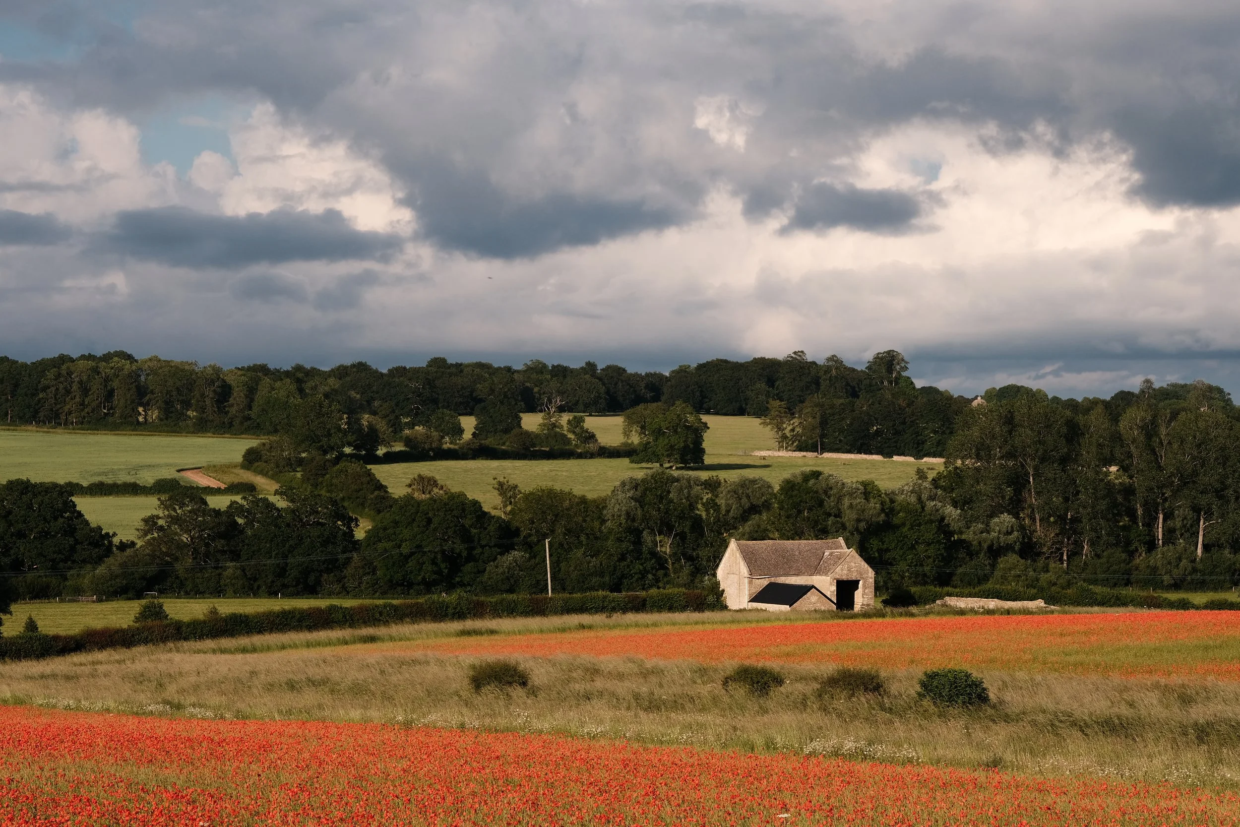 A wide Oxfordshire countryside view under a dramatic cloudy sky, with a stone barn, green fields and trees, and a red poppy field in the foreground.