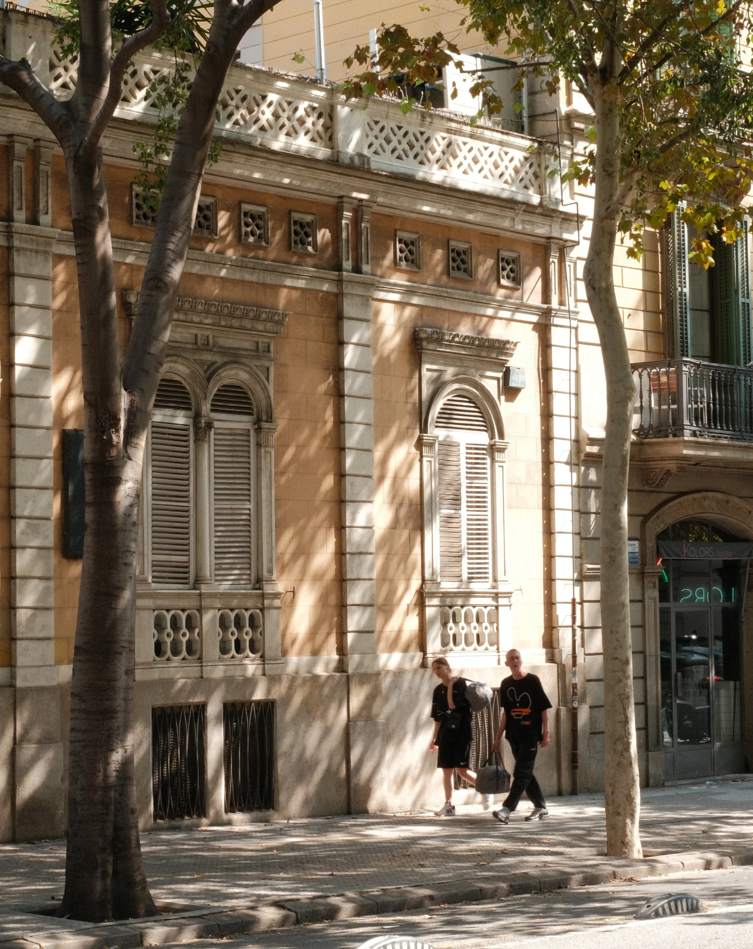 A city street scene showing a historic building with ornate windows and a balcony, two trees with thick trunks and green leaves, and two pedestrians walking along the sidewalk. One pedestrian is a woman with a backpack, and the other is a man holding
