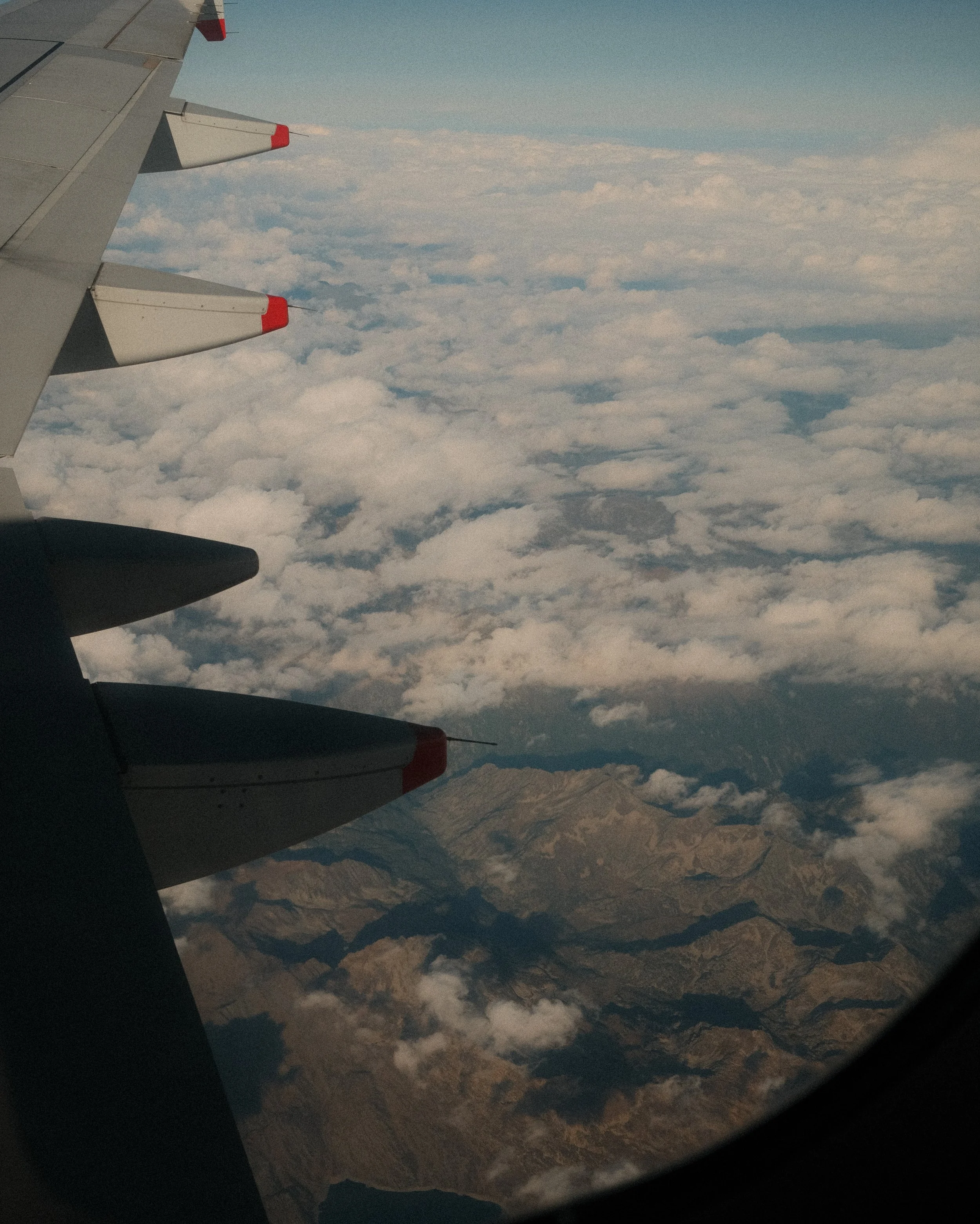 View from an airplane window showing part of the wing with red-tipped winglets and a landscape of mountains and clouds below.