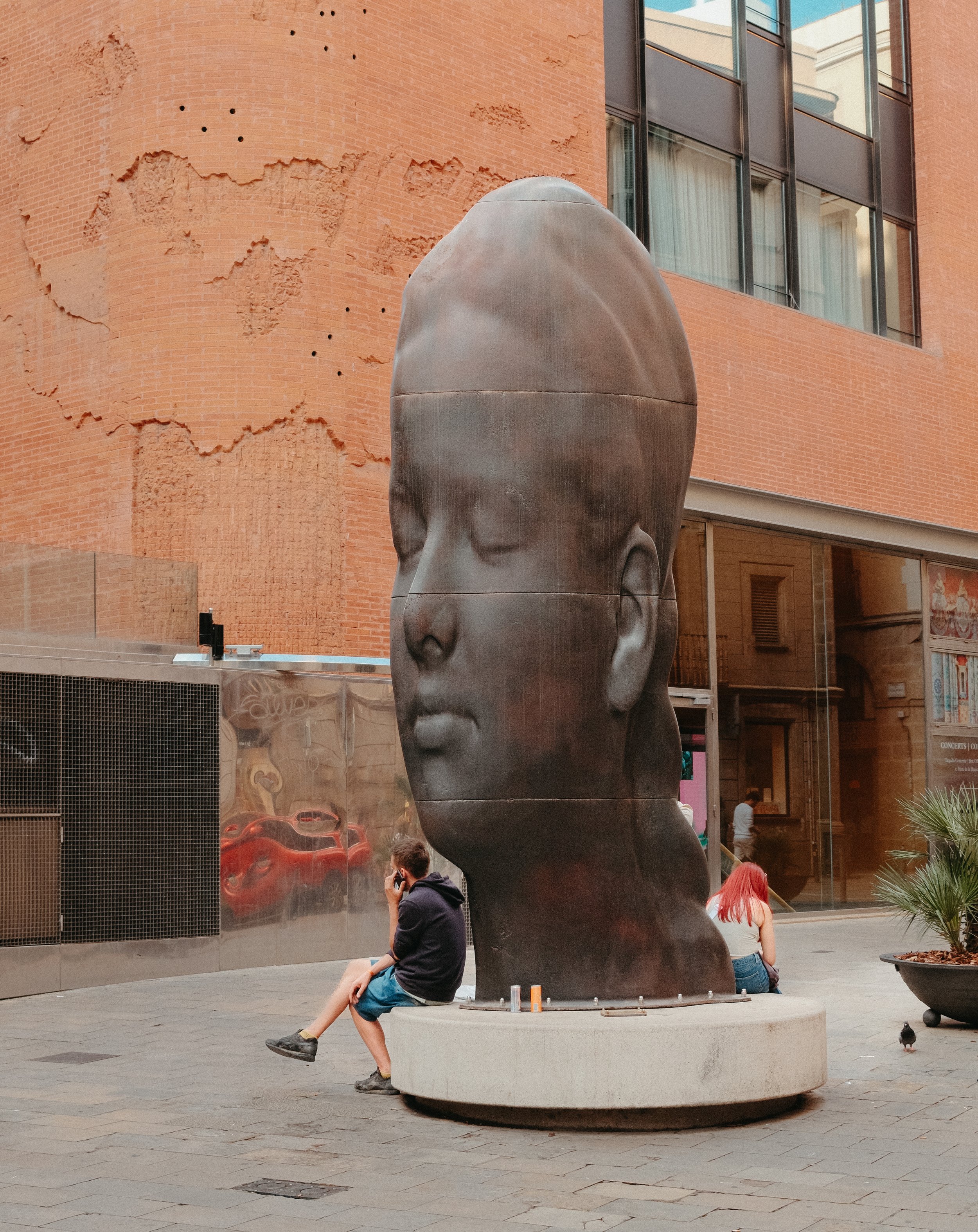 A person with red hair and a black shirt sitting on the edge of a large, abstract metal sculpture of a human head installed outdoors, with buildings and other people visible in the background.
