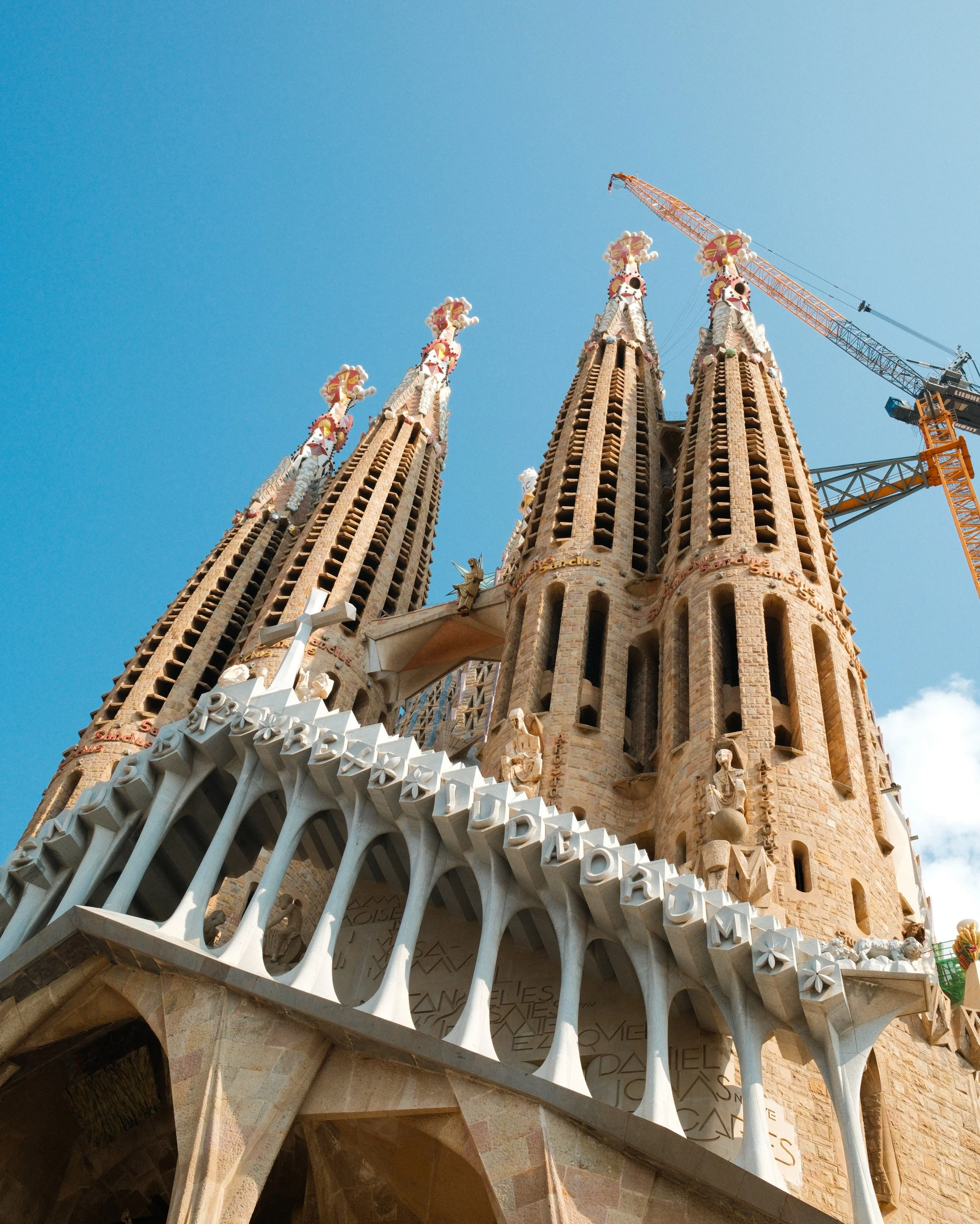Close-up view of the towers and intricate architecture of the Sagrada Família basilica in Barcelona, Spain, with a construction crane in the background against a bright blue sky.