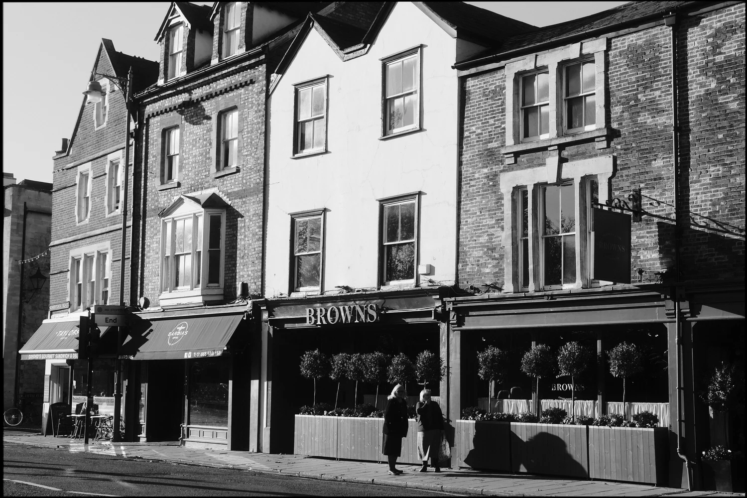 Black and white street scene outside Browns restaurant, with two people talking on the pavement and long morning shadows.
