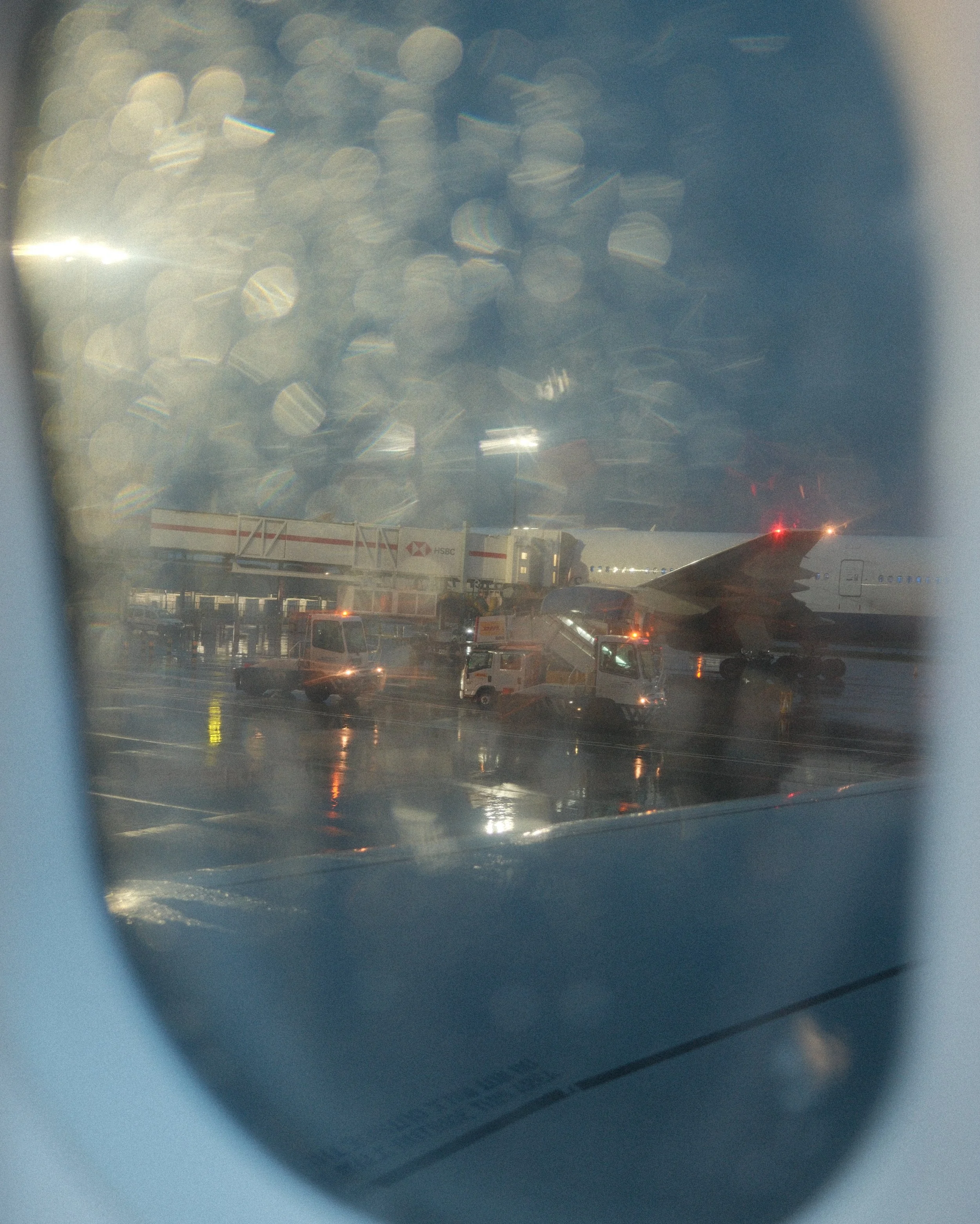 View of an airport tarmac with airplanes, vehicles, and a jet bridge in rainy weather, seen through an airplane window with raindrops on the glass.