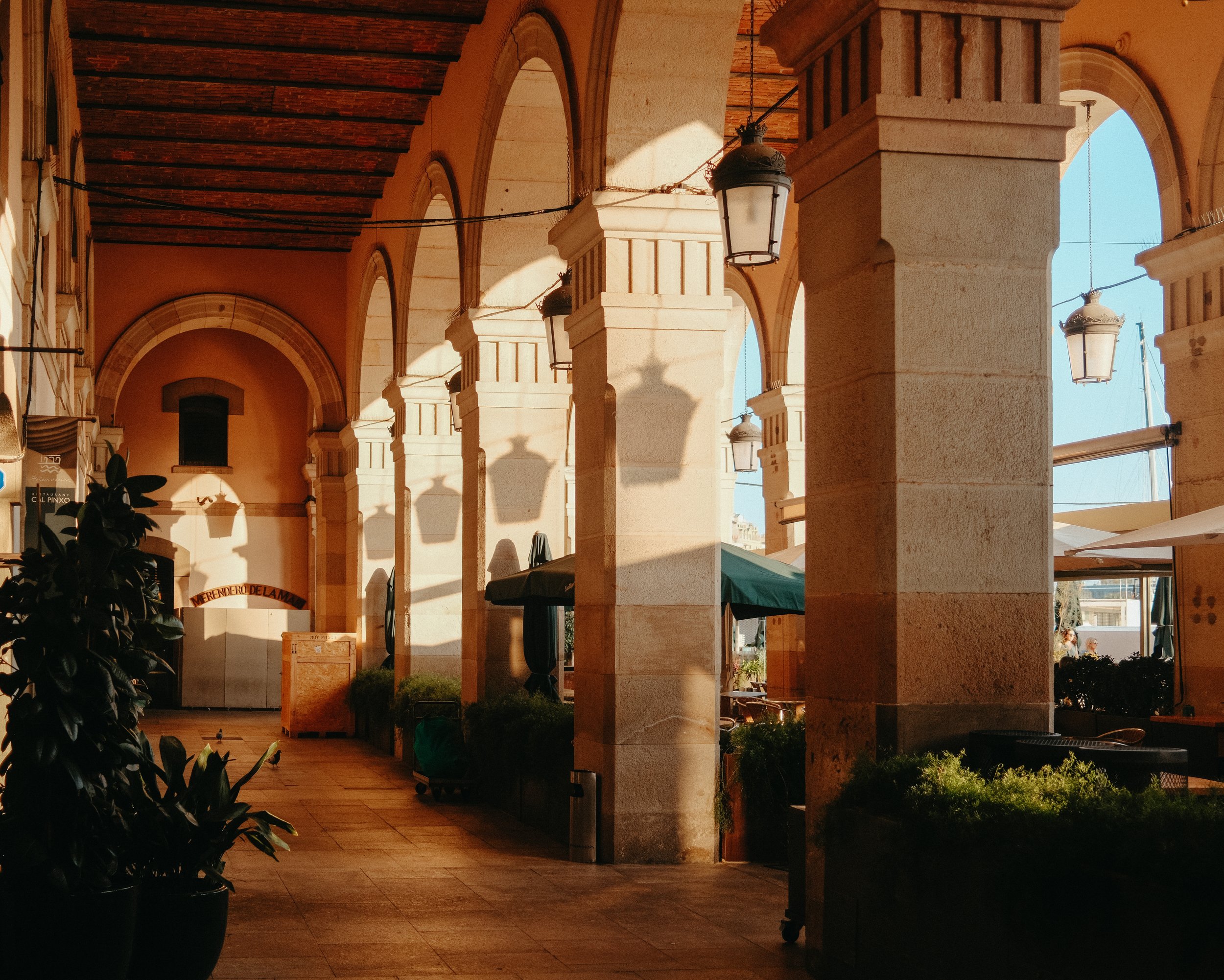 Covered outdoor patio with stone columns, hanging lanterns, and arched openings showing blue sky and part of a building in the background.
