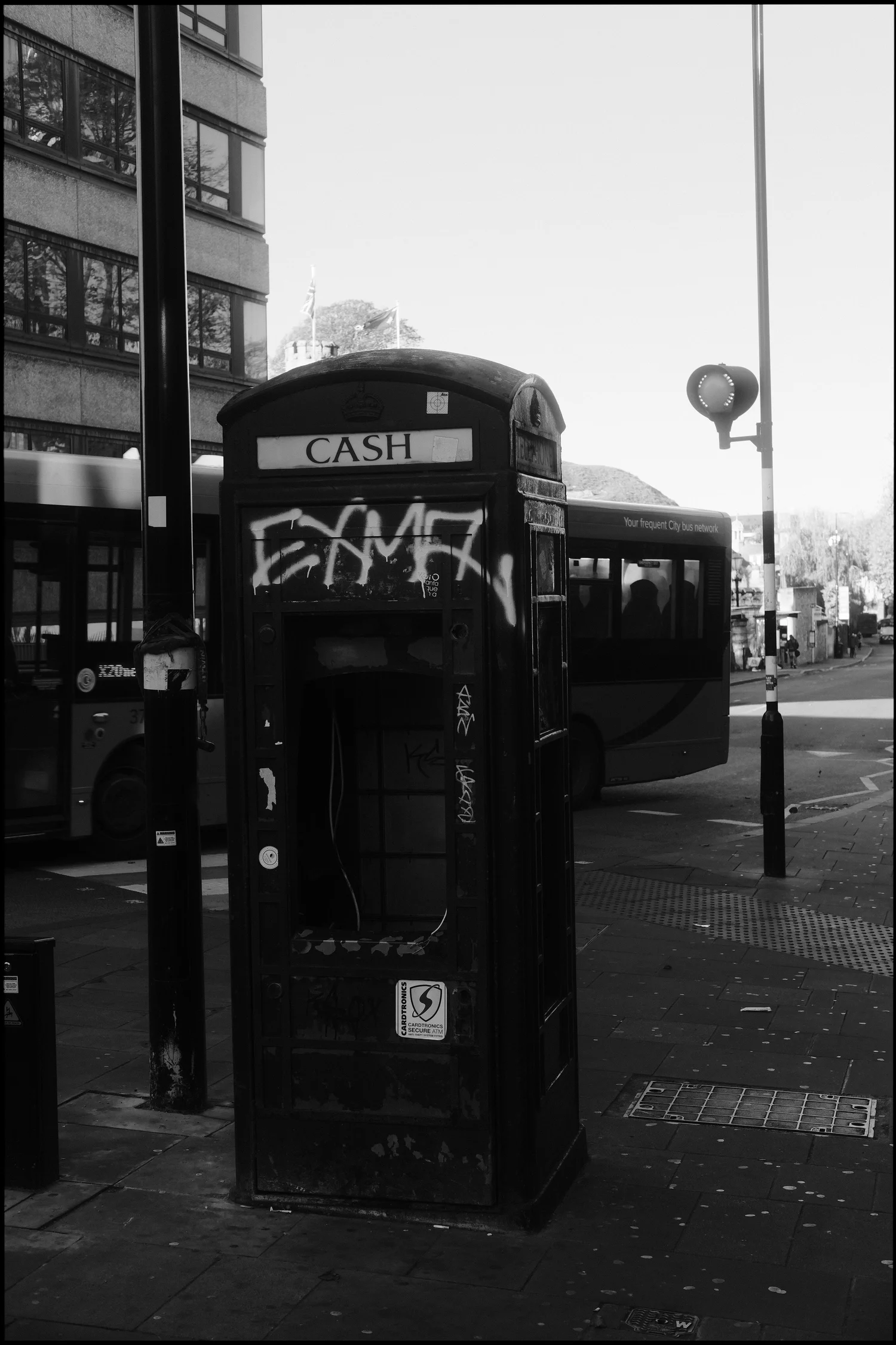 Black and white photo of a gutted, graffitied phone box on a street corner with a bus blurred in motion behind it.