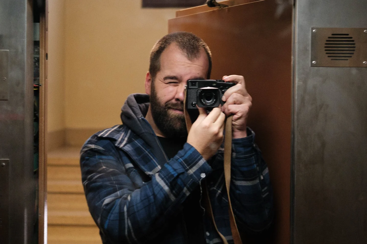 Man taking a selfie in an elevator mirror, wearing a plaid jacket and holding camera.