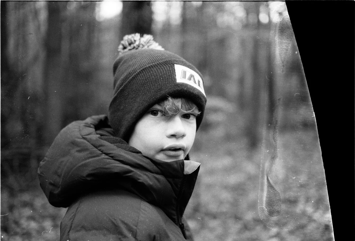 A boy in a beanie hat looks back warily over his shoulder in the woods.