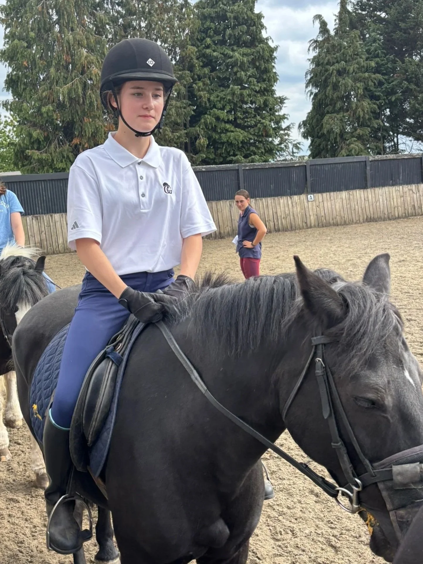 A young person in a white polo shirt, navy riding pants, black gloves, and a black riding helmet sits on a black horse, looking straight ahead with a serious expression. Other people and trees are visible in the background at an outdoor riding arena with a sandy surface.