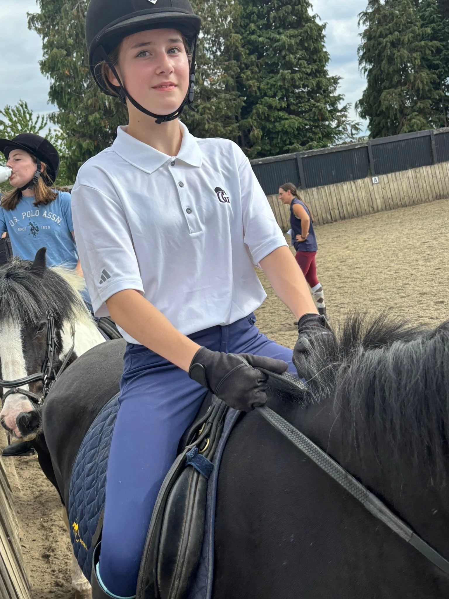 Young person wearing a white polo shirt, black helmet, and black gloves riding a black horse during a daytime equestrian activity.