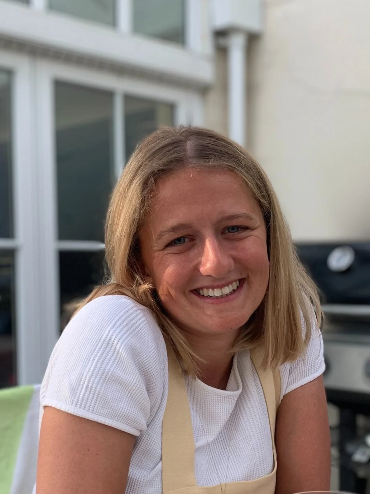 A person smiling while sitting outside near a building with large windows and a grill in the background.