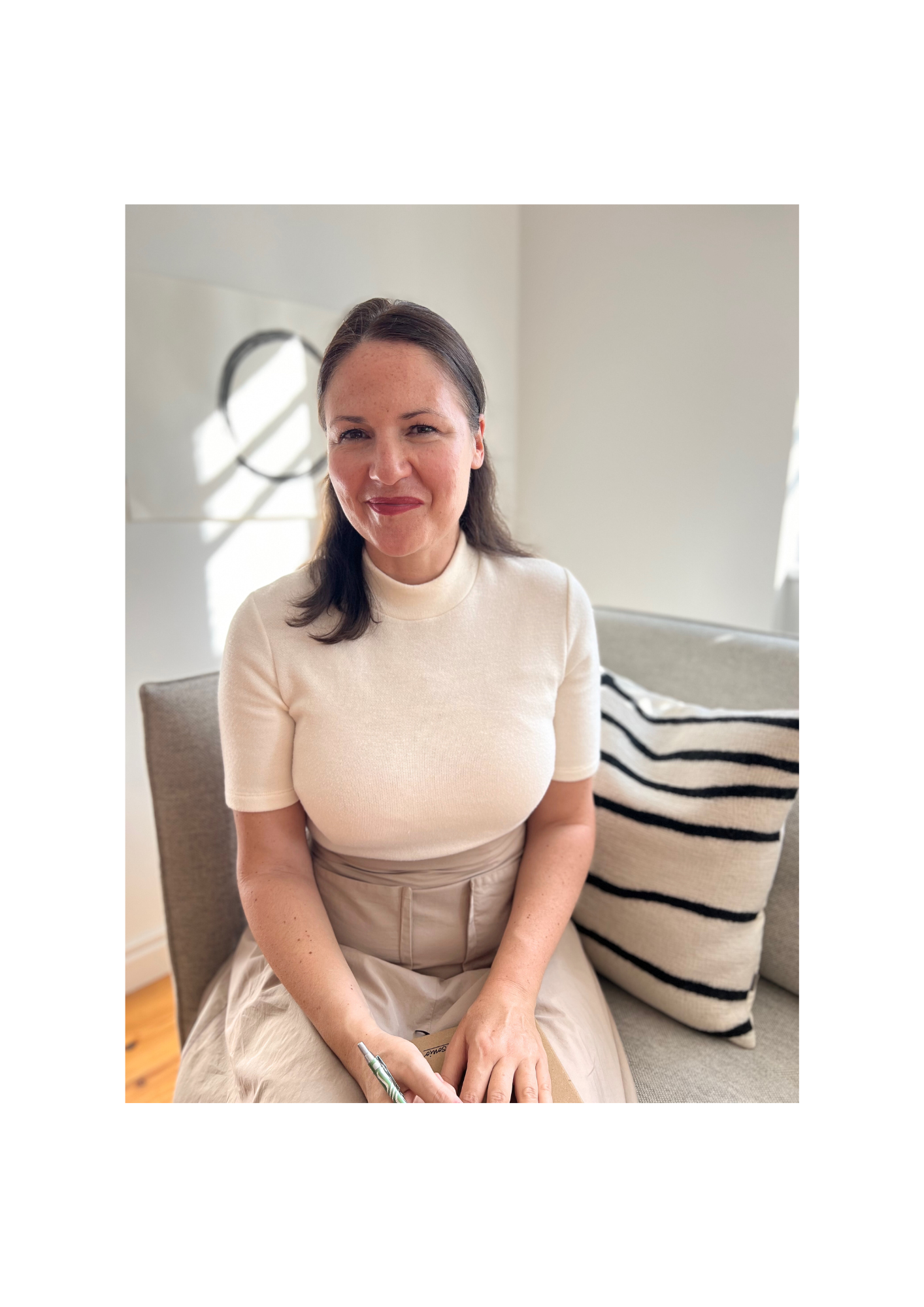 Eine Heilpraktikerin für Psychotherapie sitzt auf einem Sofa in einem hell erleuchteten Raum, trägt ein cremefarbenes-beiges Kleid und schaut in die Kamera.