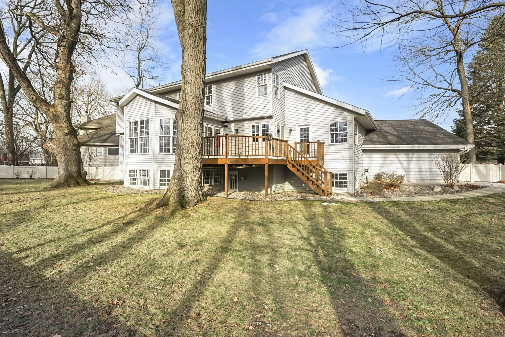 A large two-story house with gray siding and a wooden deck in the backyard, trees without leaves, a grassy lawn, and a blue sky with some clouds.