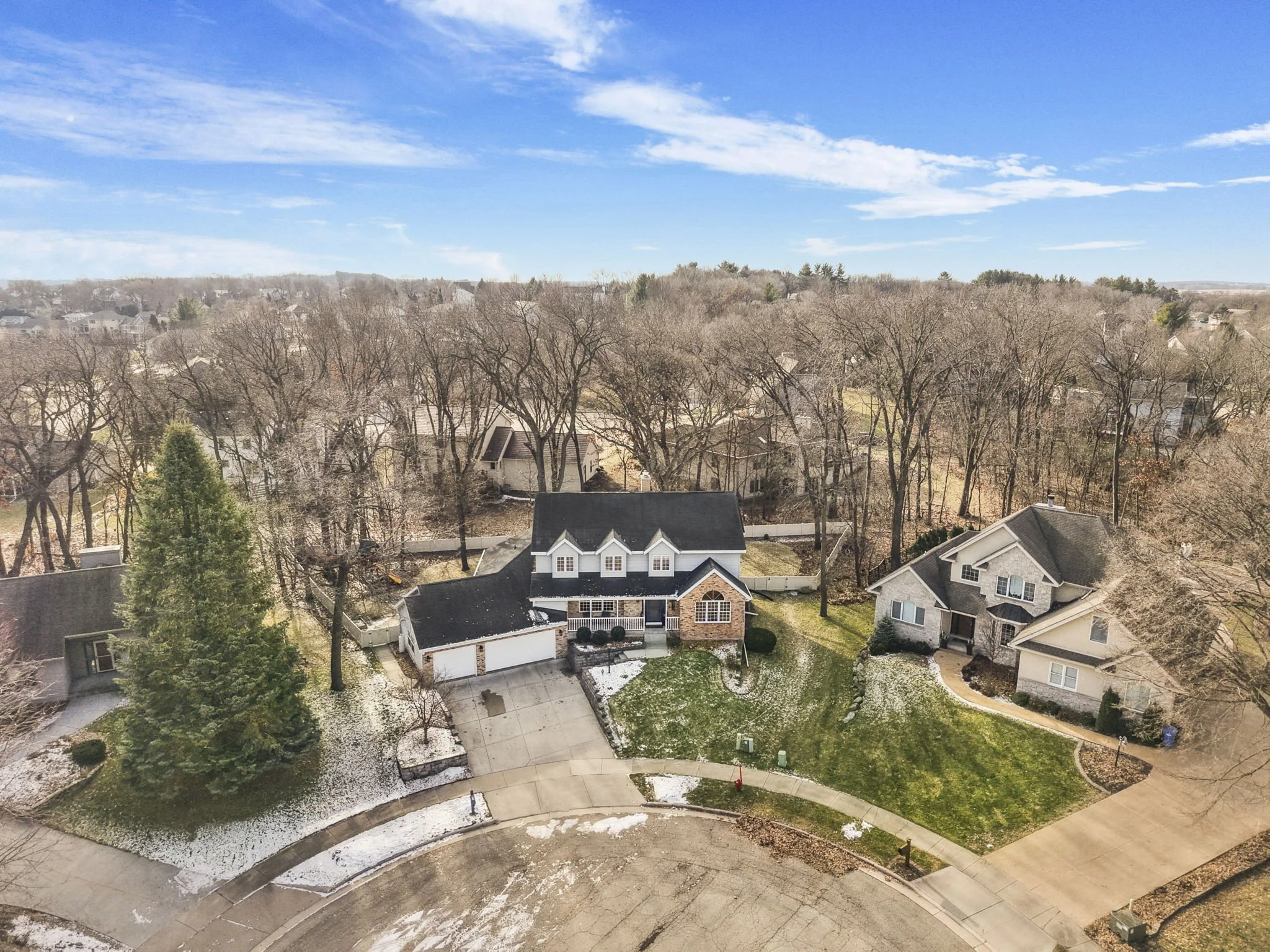 An aerial view of suburban houses with trees, a curved sidewalk, and a clear blue sky.